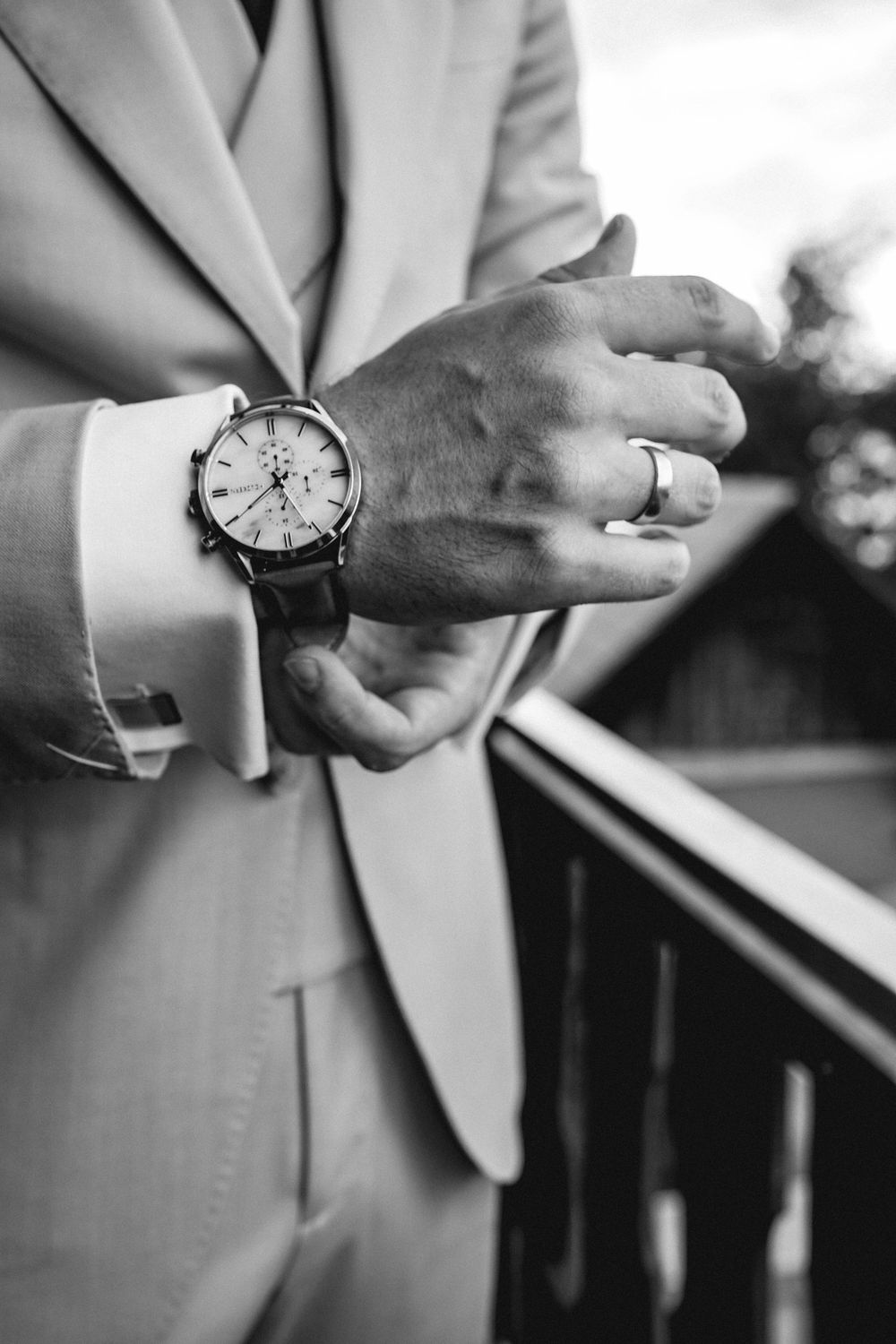 Close-up black and white photo of a wrist wearing an elegant watch and wedding ring with a light colored suit sleeve.