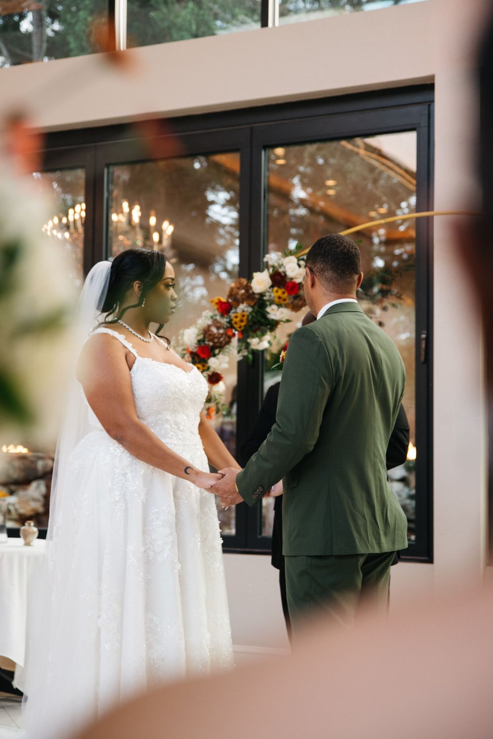 Couple holds hands during intimate wedding ceremony in front of floral-decorated windows.