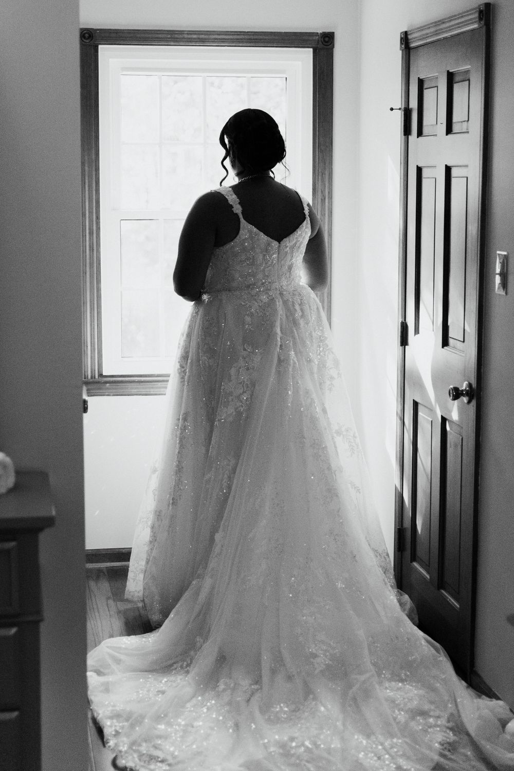 Silhouette of bride in lace wedding dress with long train standing by window.