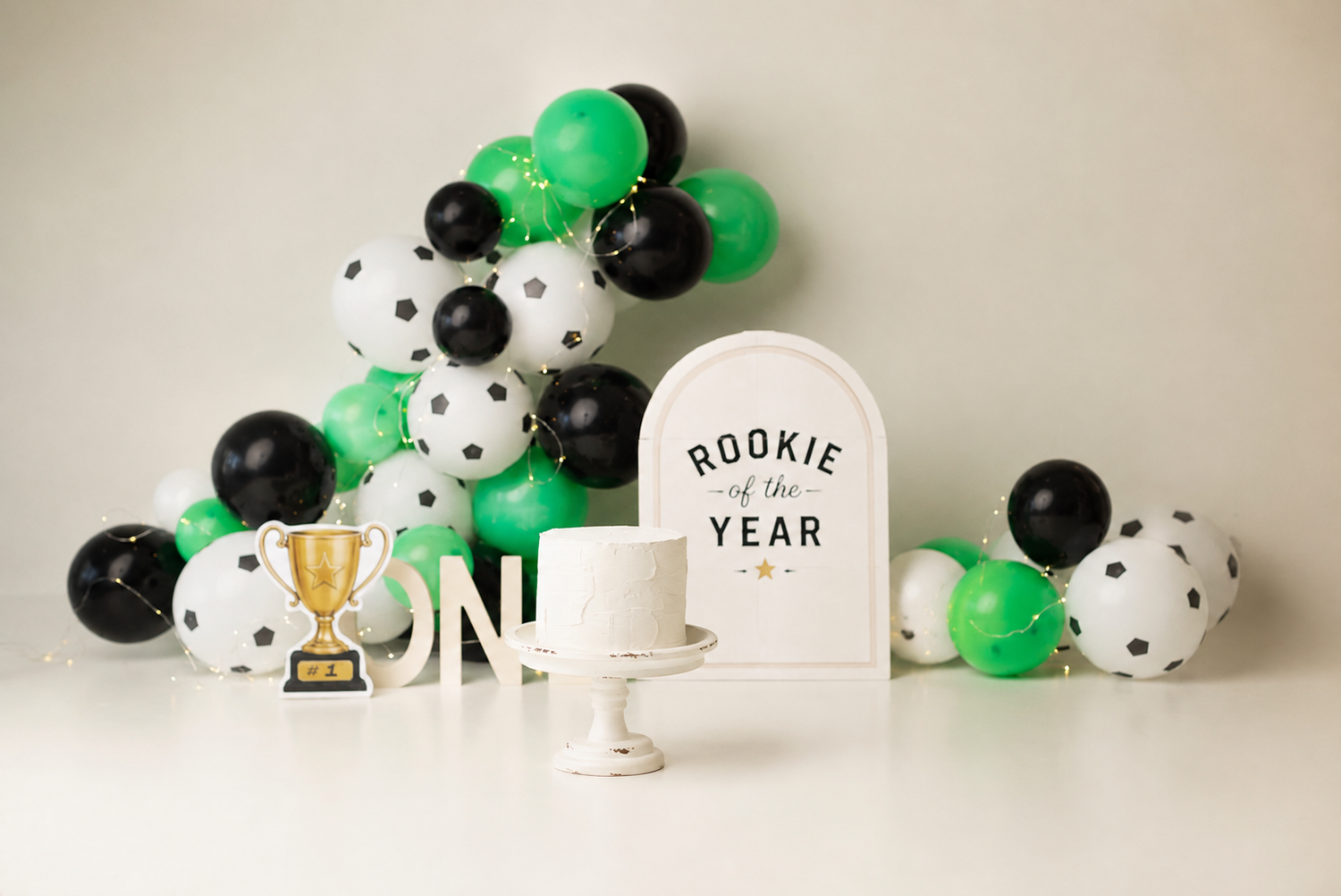 Soccer-themed cake smash backdrop with green, black, white balloons, Rookie of the Year sign, trophy, and white cake on stand.