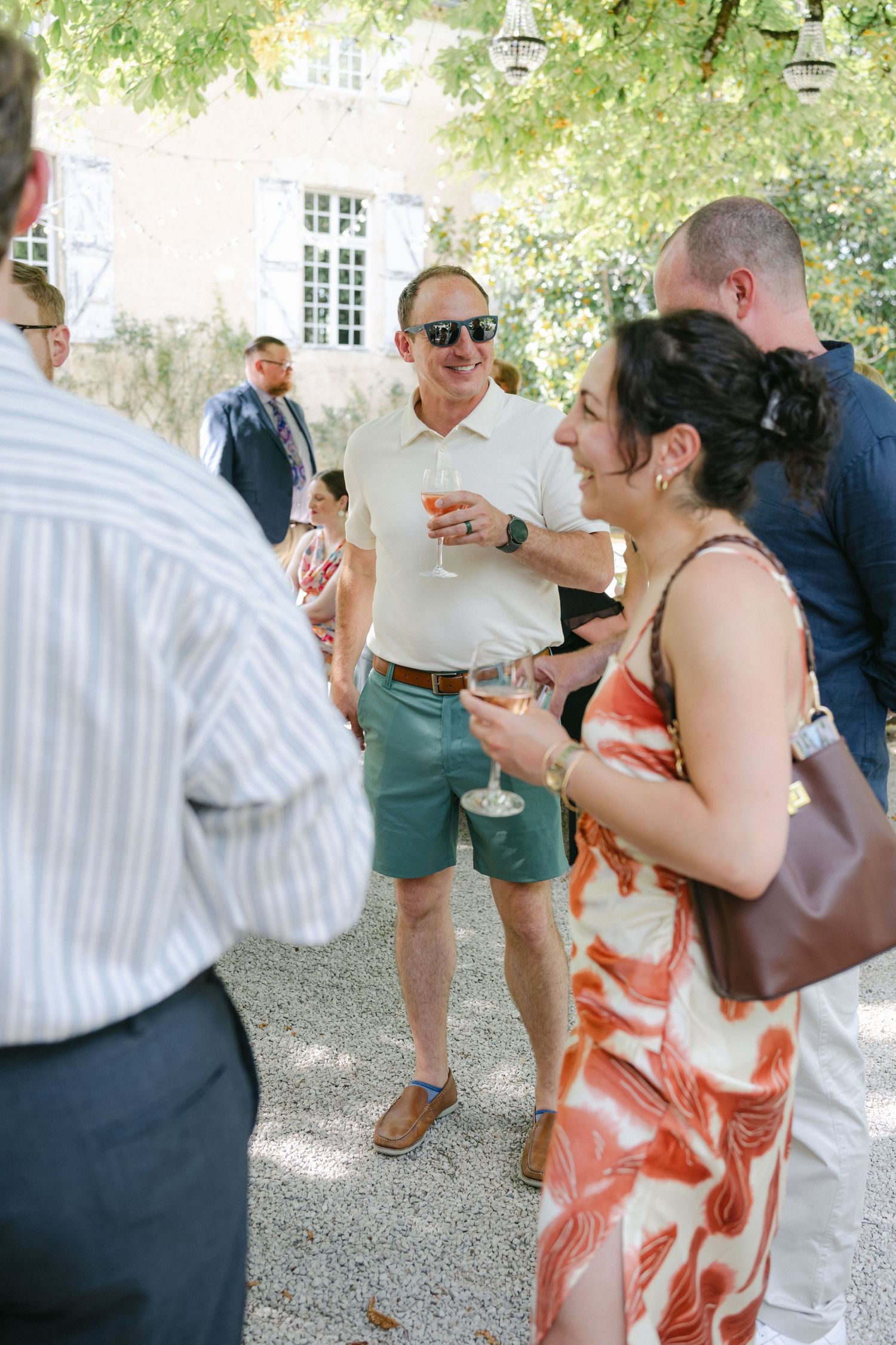 Guests in summer attire socializing outdoors at a garden party, holding champagne glasses near a historic building.