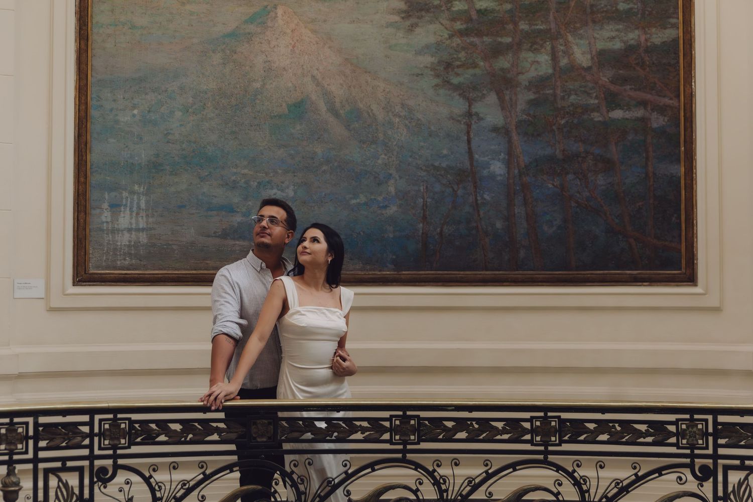 Couple gazing upward together on a decorative balcony beside a large oil painting.