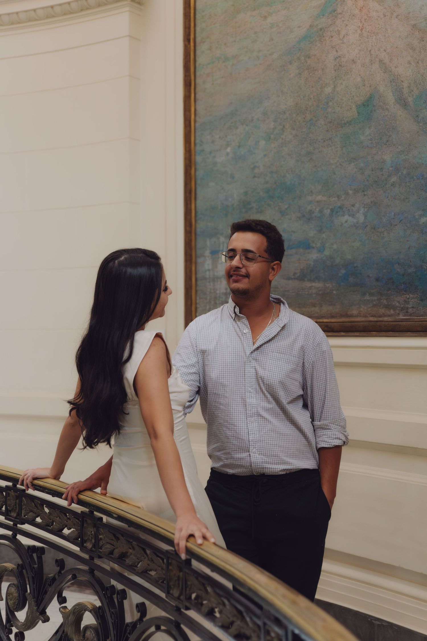 Couple holding hands and smiling on a decorative staircase with a large painting behind.