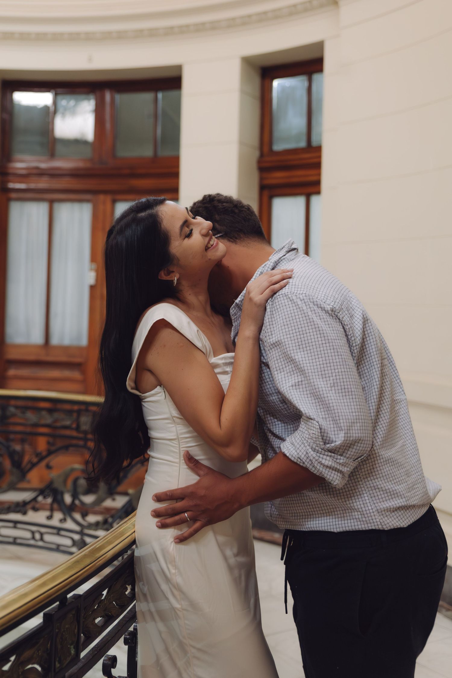 Couple sharing a romantic kiss on a balcony railing inside an elegant historic building.