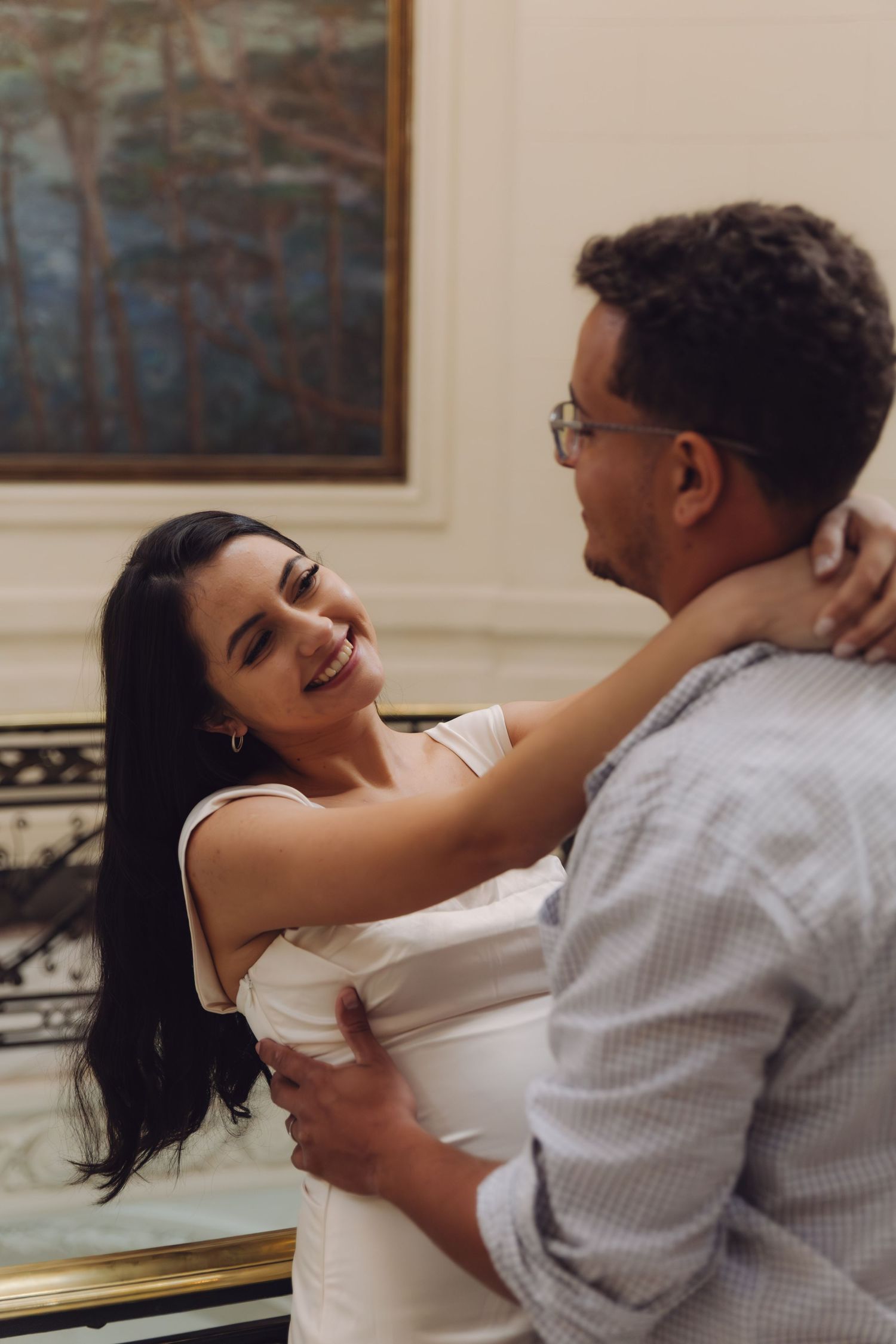 A smiling woman in a white dress embraces a man wearing glasses inside an elegant room with classic paintings.