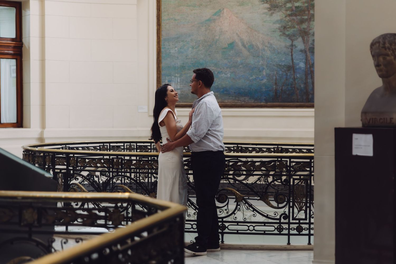 A couple shares a romantic embrace on a balcony inside a grand building with a mountain landscape painting behind them.