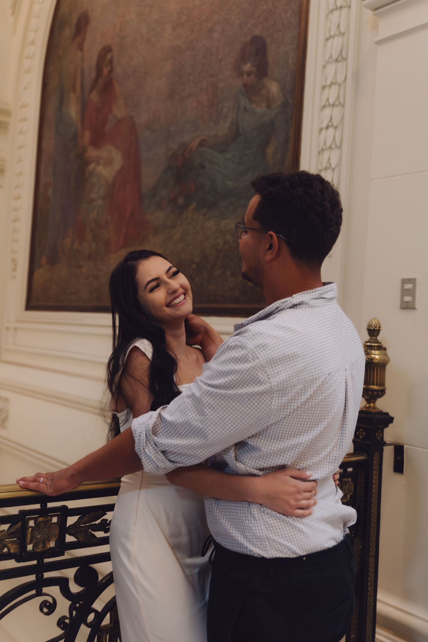 A joyful couple embraces near an ornate railing with a classical painting backdrop in an elegant interior.