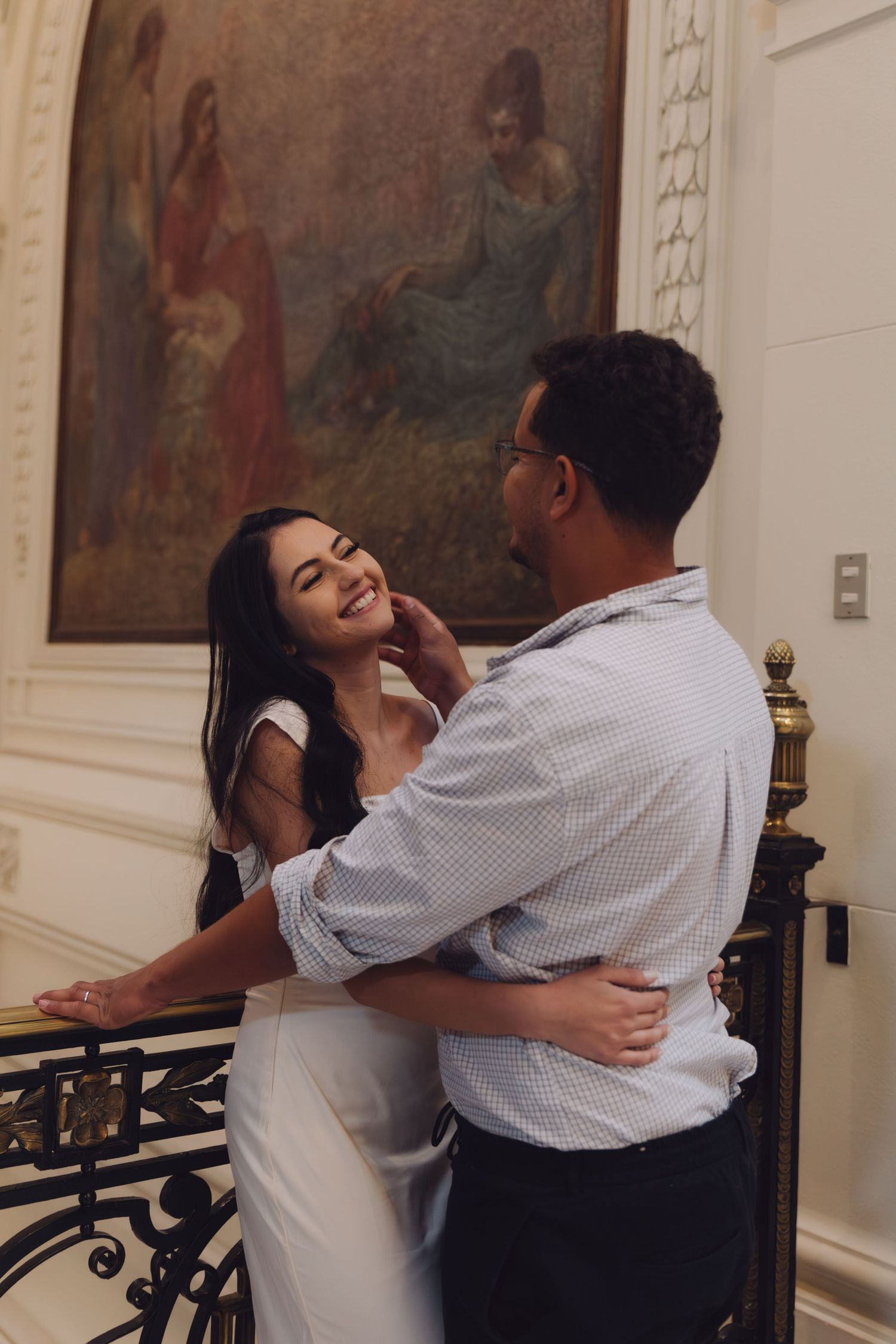 A man tenderly cups a laughing woman's face as they embrace near a decorative railing in a classic interior.