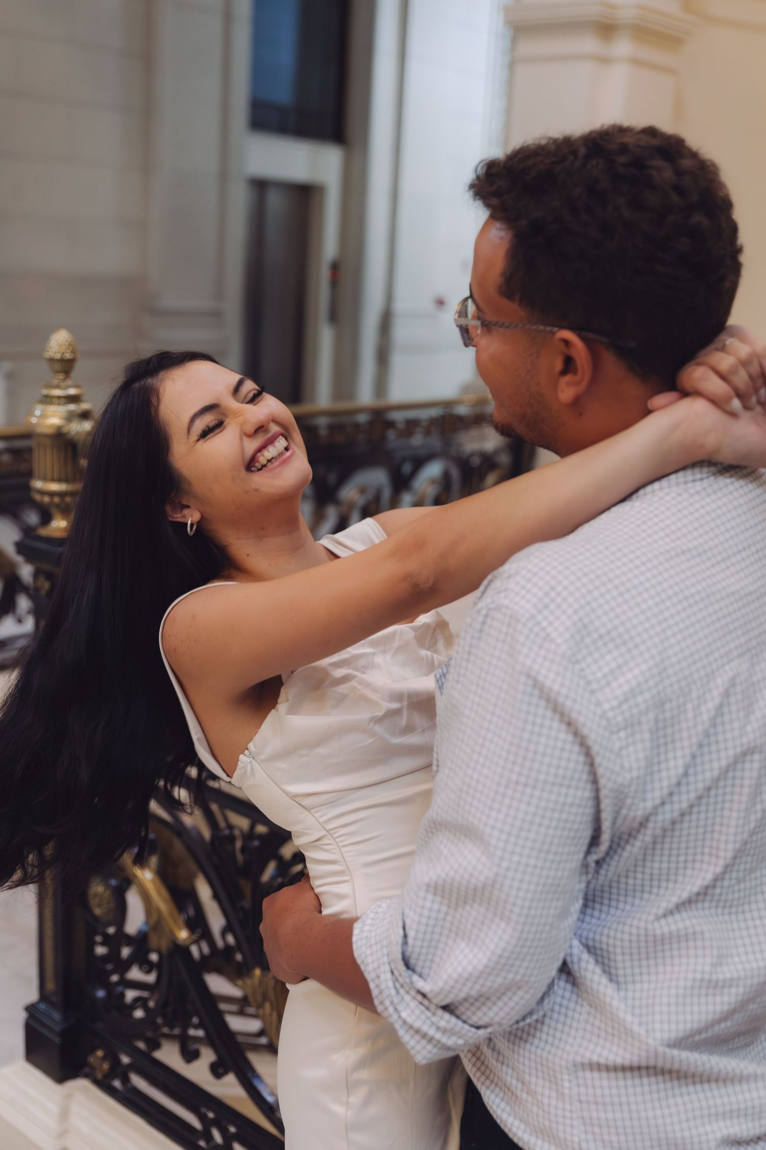 A laughing woman in a white dress wraps her arms around a man near an ornate staircase railing.