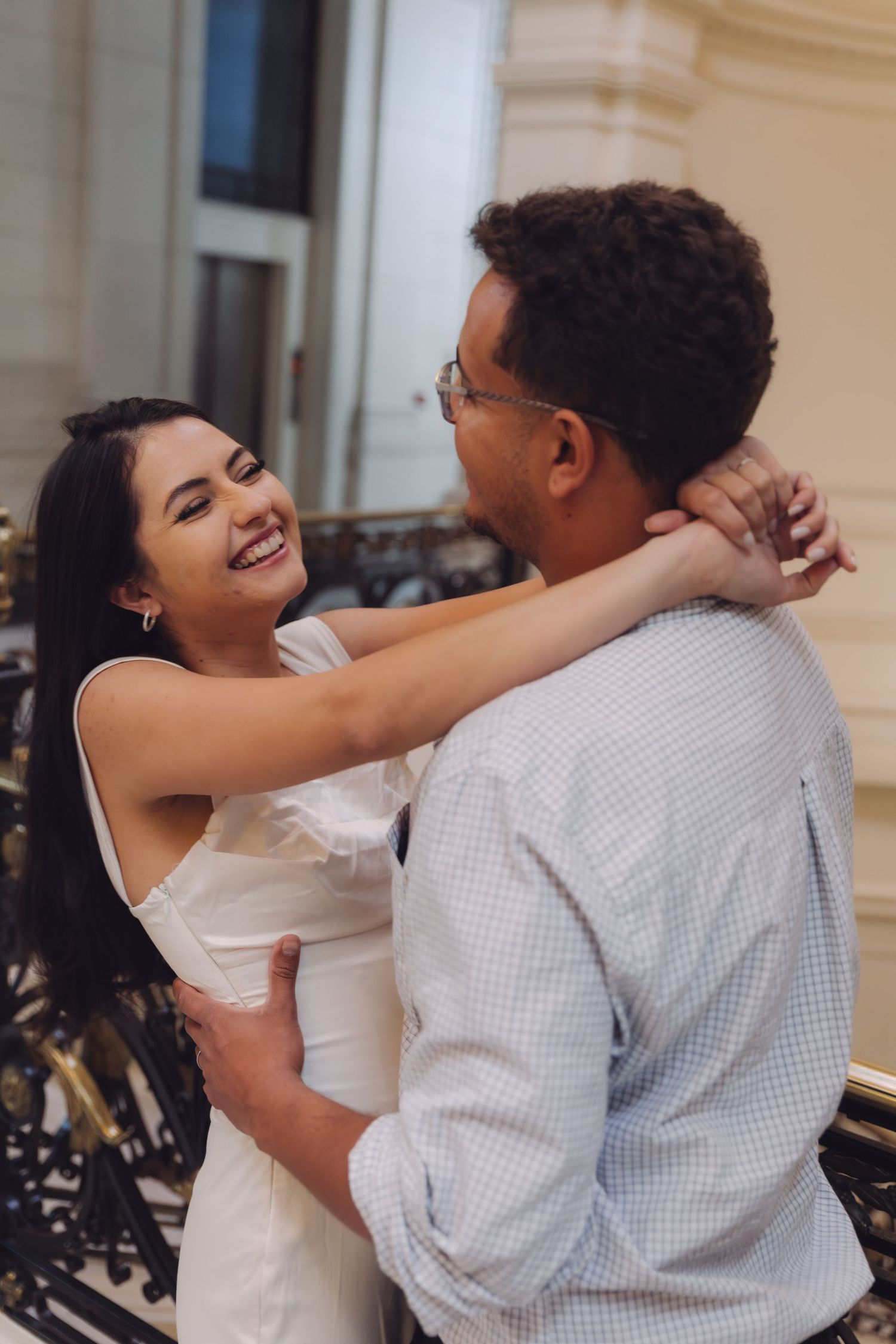 A smiling woman in a white dress embraces a man wearing glasses near an elegant black wrought-iron railing.