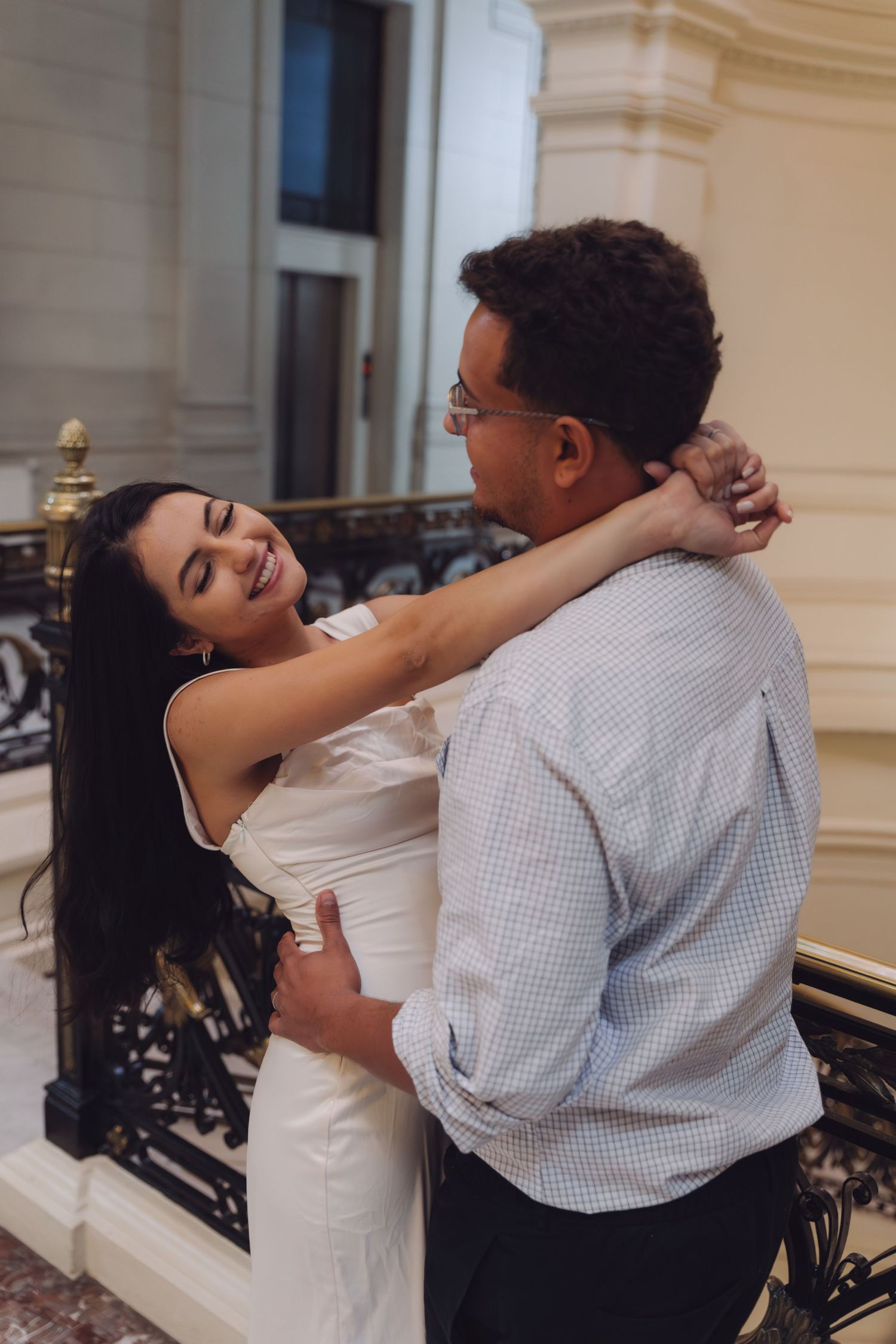 A happy couple shares a warm embrace near an ornate black railing in a grand classical interior setting.