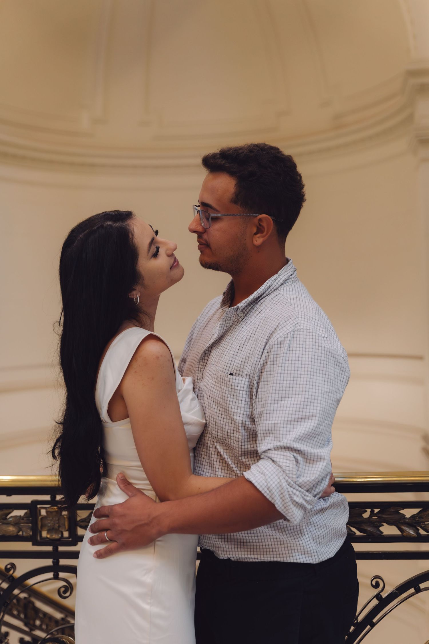 Couple sharing an intimate moment, embracing on an ornate balcony in an elegant white interior.