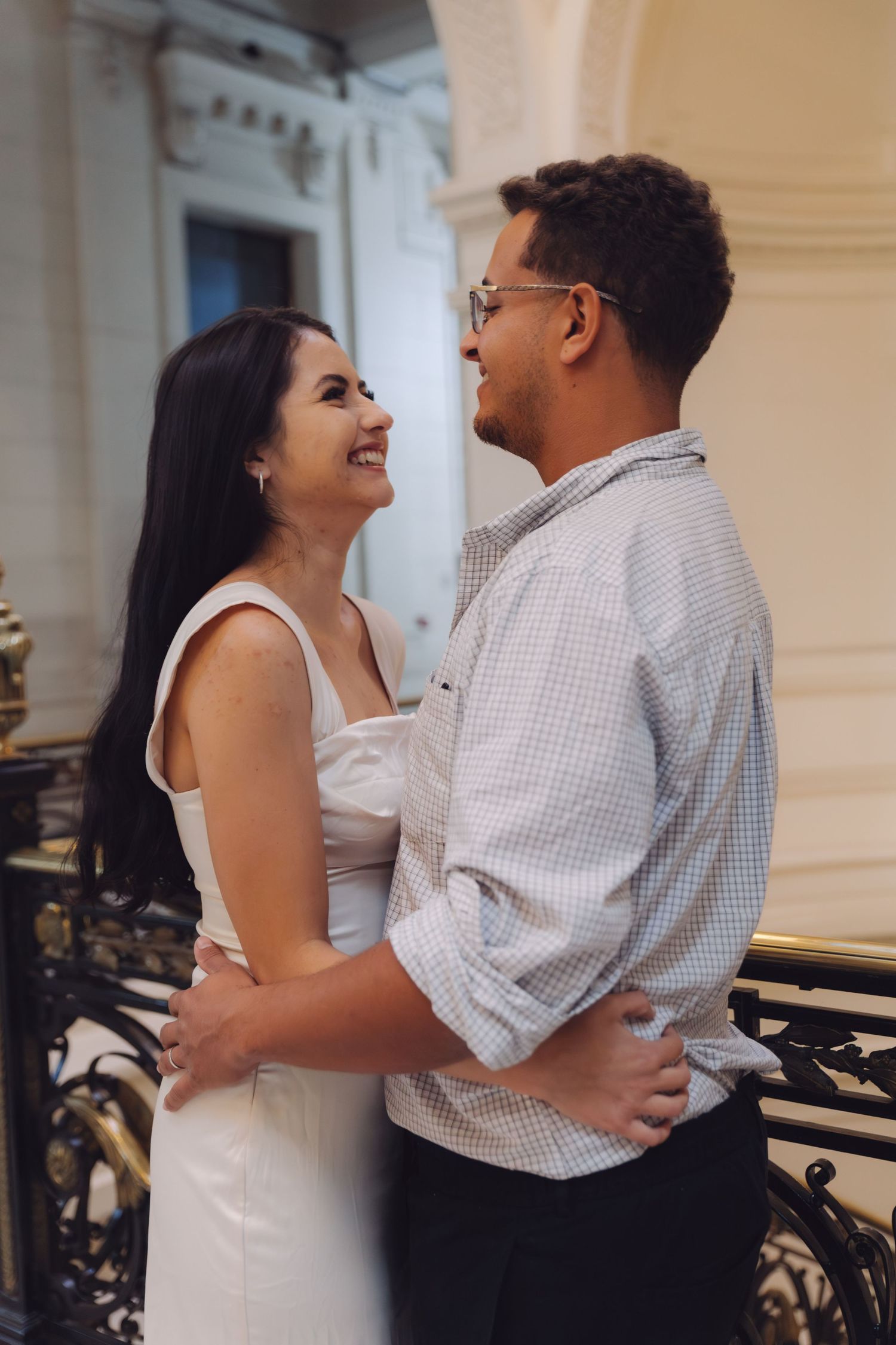 Smiling couple embracing and gazing at each other on a decorative iron railing balcony.