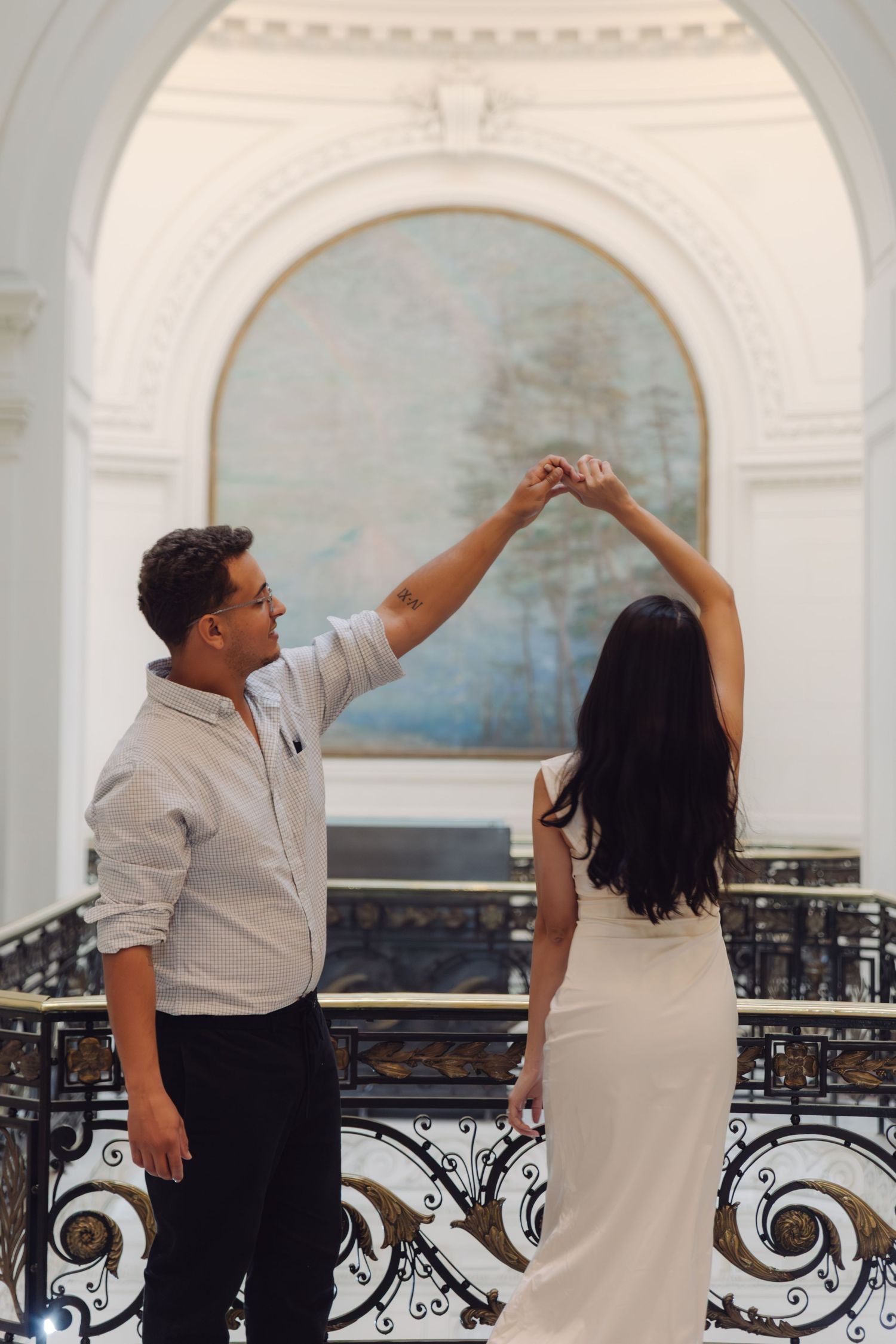 Couple dancing gracefully in an elegant white hall with ornate railings and arched artwork.