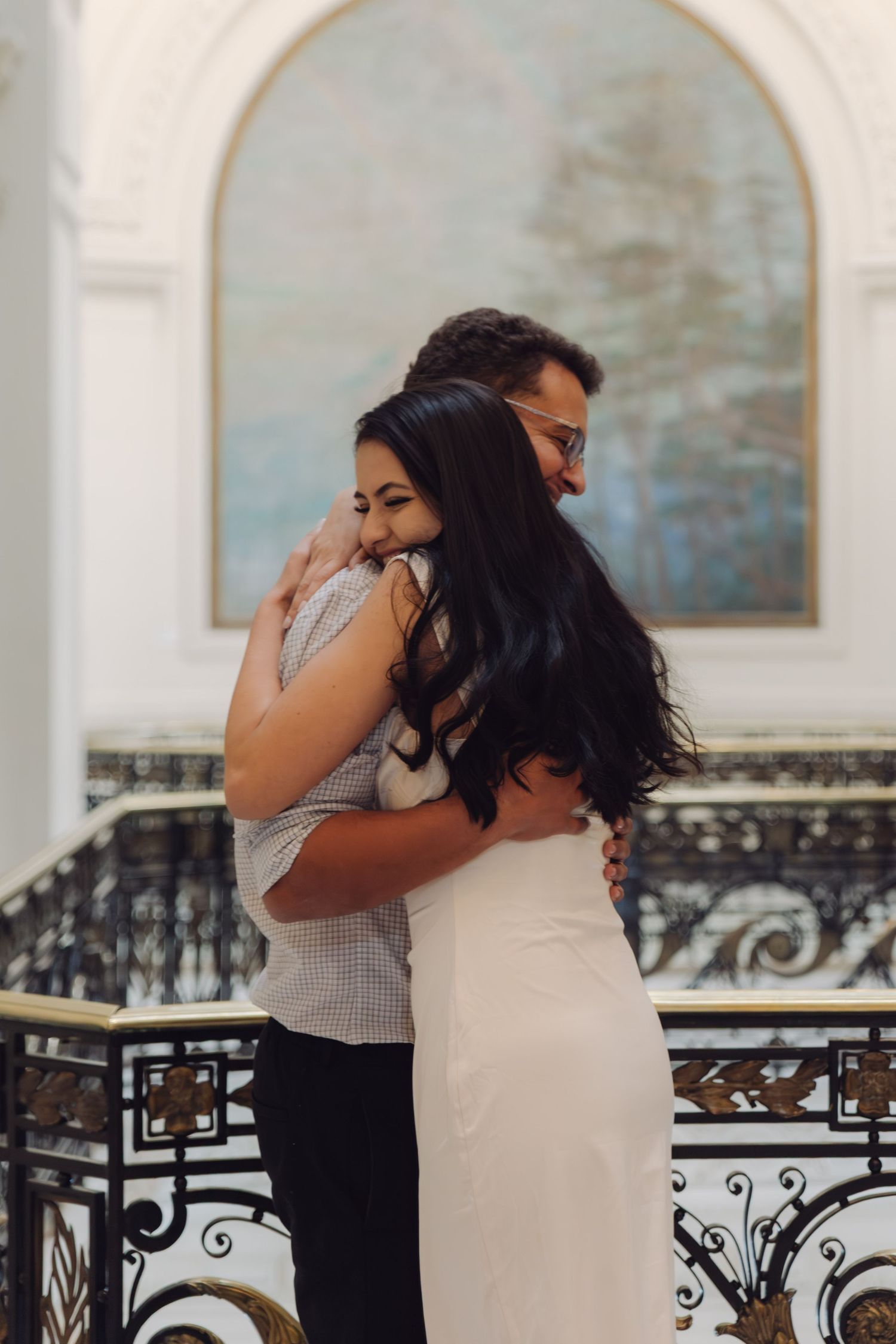 Couple sharing a warm embrace in front of an arched painting in a grand interior.