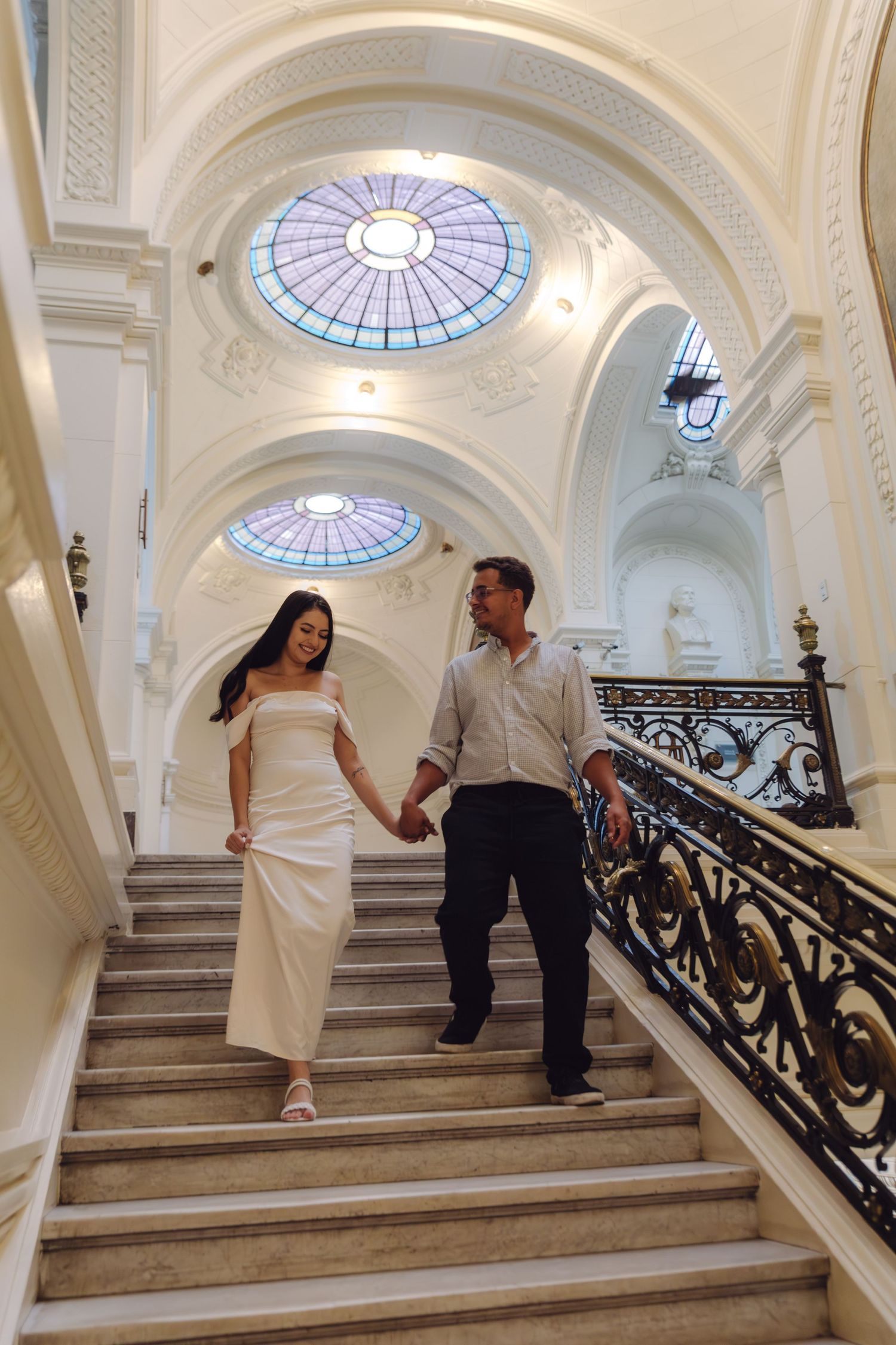 Couple holding hands while descending a grand marble staircase beneath a stained glass dome.