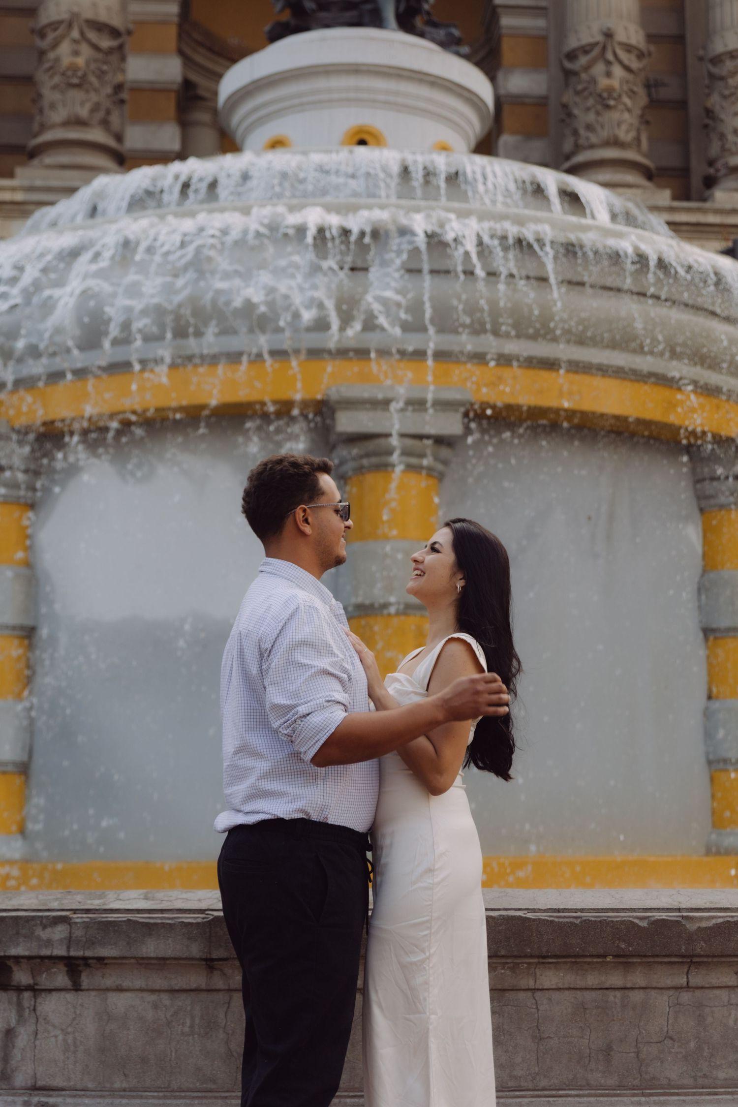 Romantic couple embracing in front of an ornate yellow and white decorative fountain outdoors.
