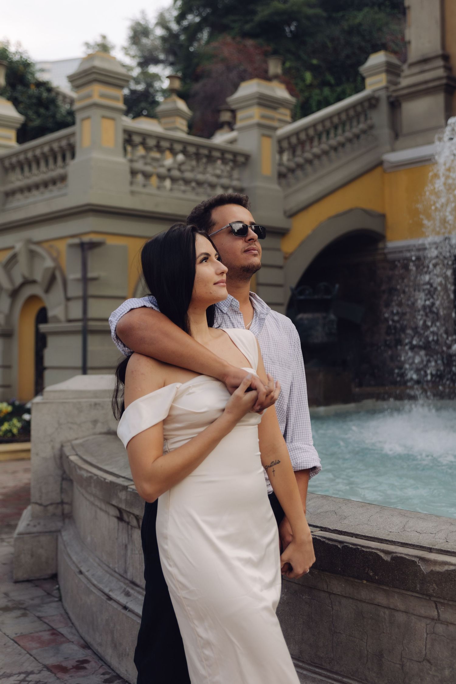 Romantic couple embracing by an ornate fountain, woman in white dress, man in sunglasses holding her close.