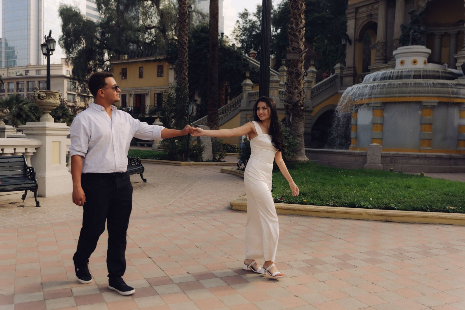 Couple playfully holding hands and dancing outdoors near a fountain surrounded by palm trees.