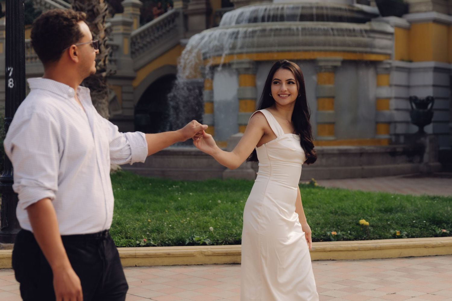 Couple dancing and holding hands near a decorative fountain, woman in elegant white gown smiling joyfully.