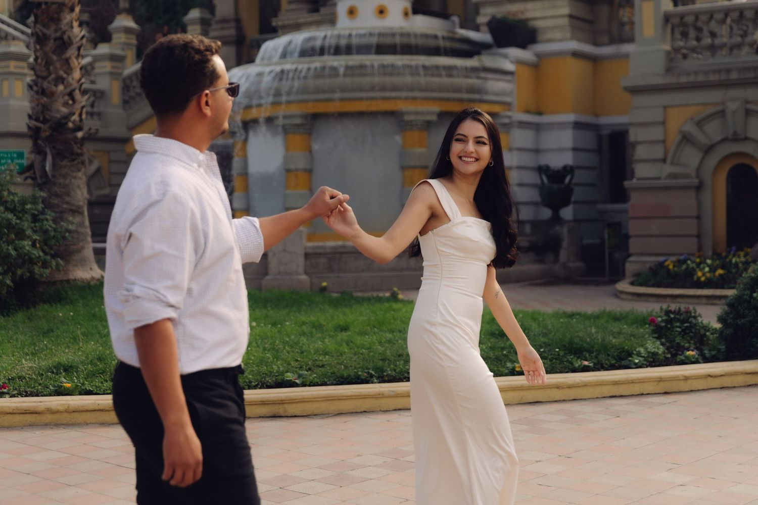 Couple holding hands and dancing near an ornate fountain, woman in white dress laughing with joy.