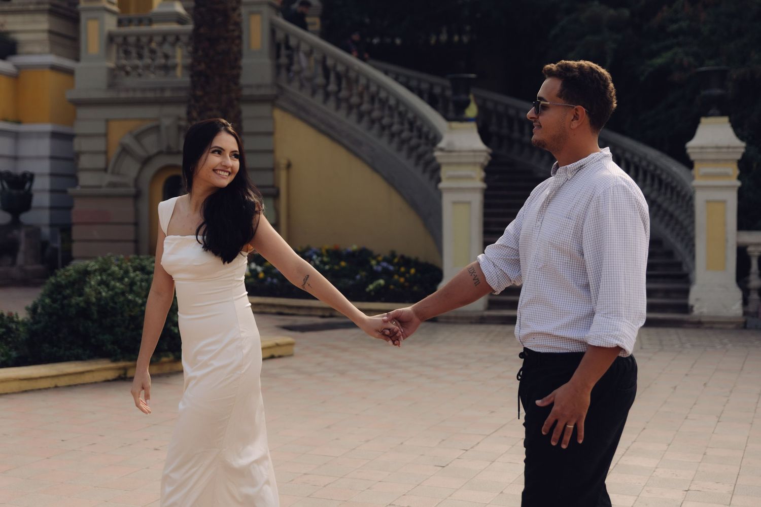 Couple smiling and holding hands while walking near a yellow ornate building and fountain.
