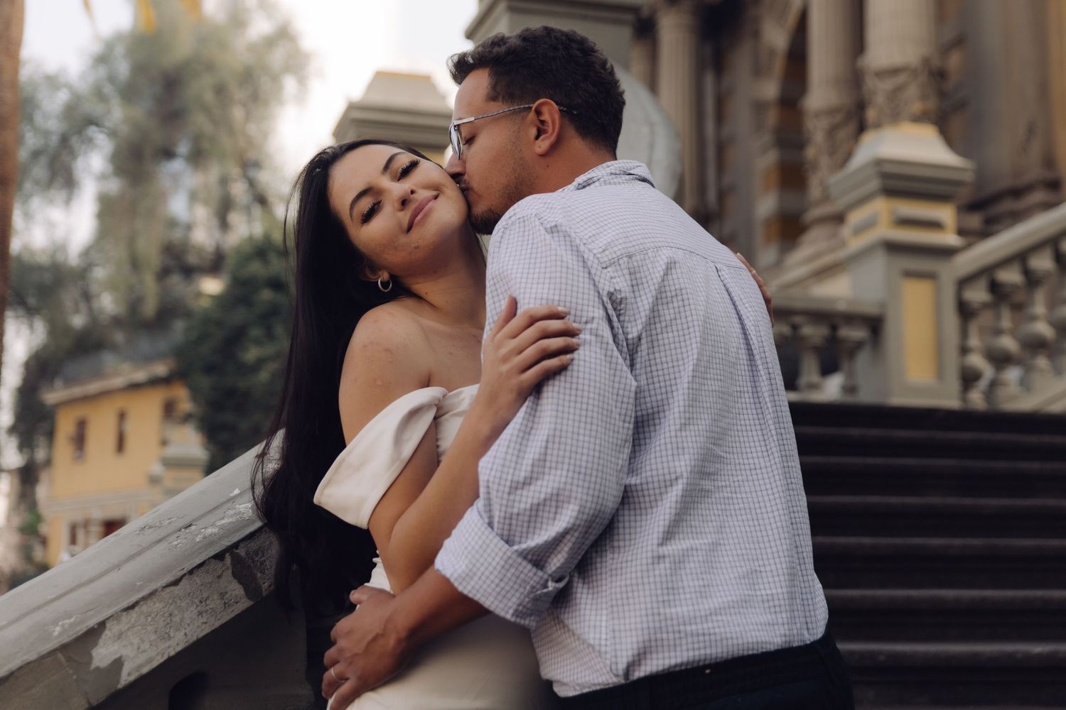 Man tenderly kissing woman on cheek while embracing on stone steps of a classic building.