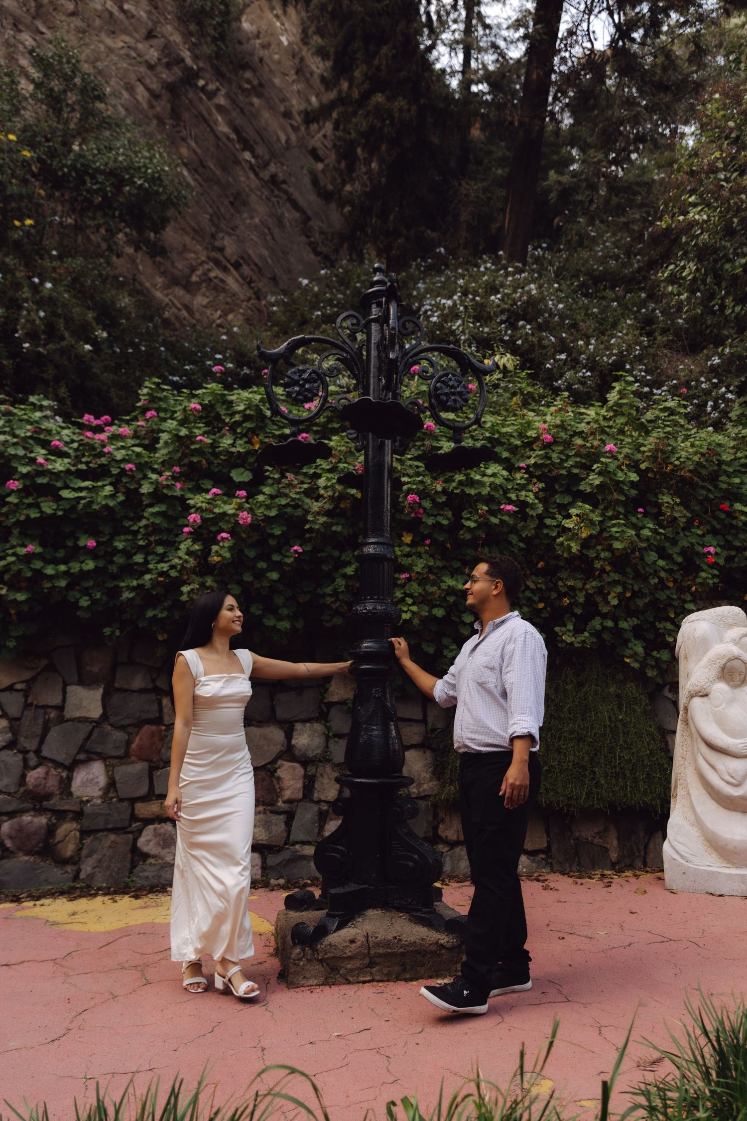 Couple posing beside an ornate black iron lamppost surrounded by flowering garden and stone wall.