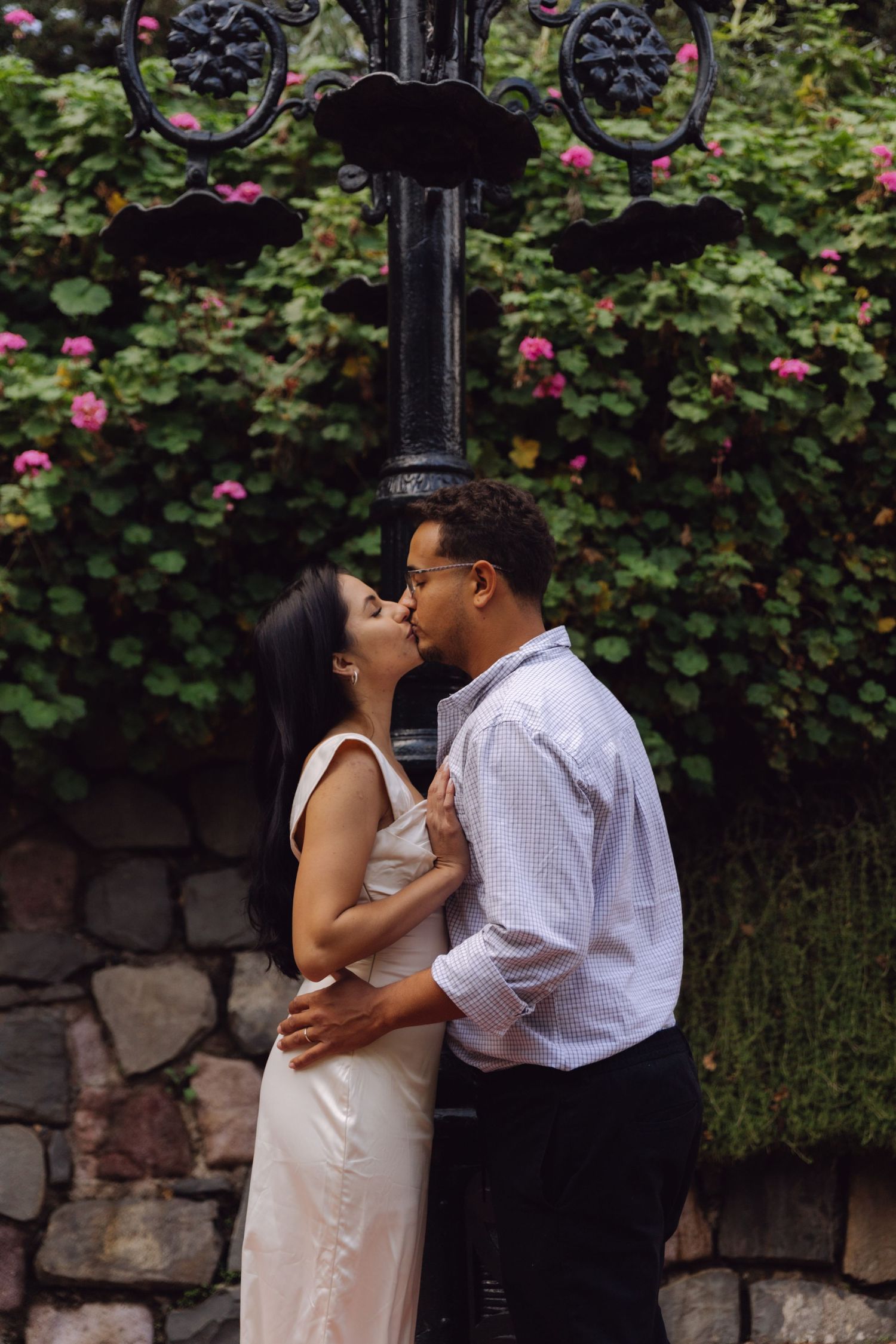 Couple sharing a romantic kiss beside a decorative iron lamppost with pink flowers in background.