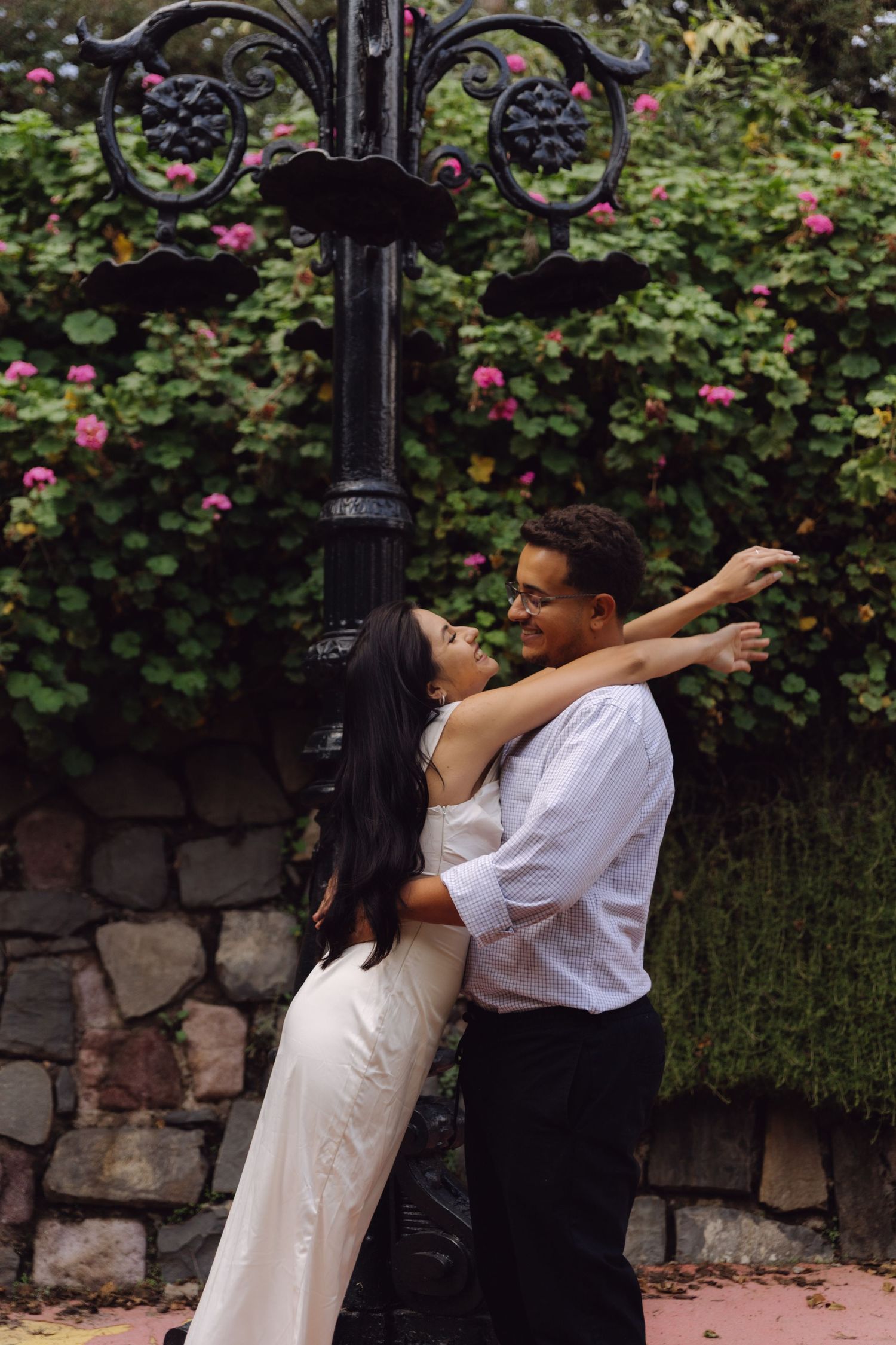 Couple embracing near ornate black lamppost with pink flowering vines on stone wall backdrop.