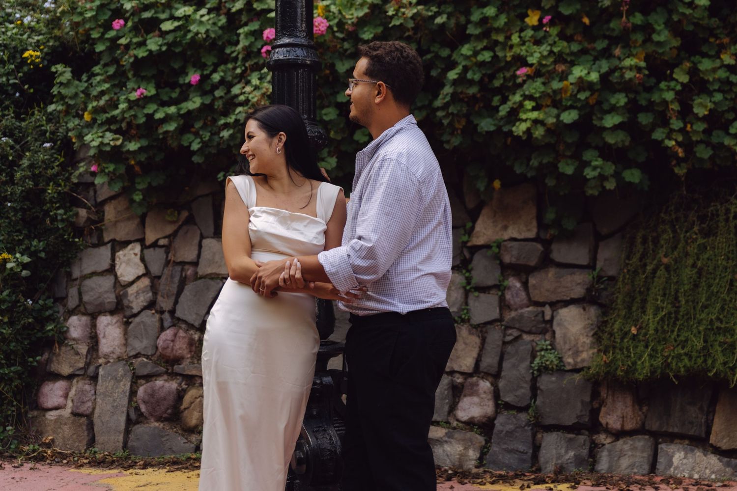 Elegant couple holding hands by black lamppost against rustic stone wall with roses.