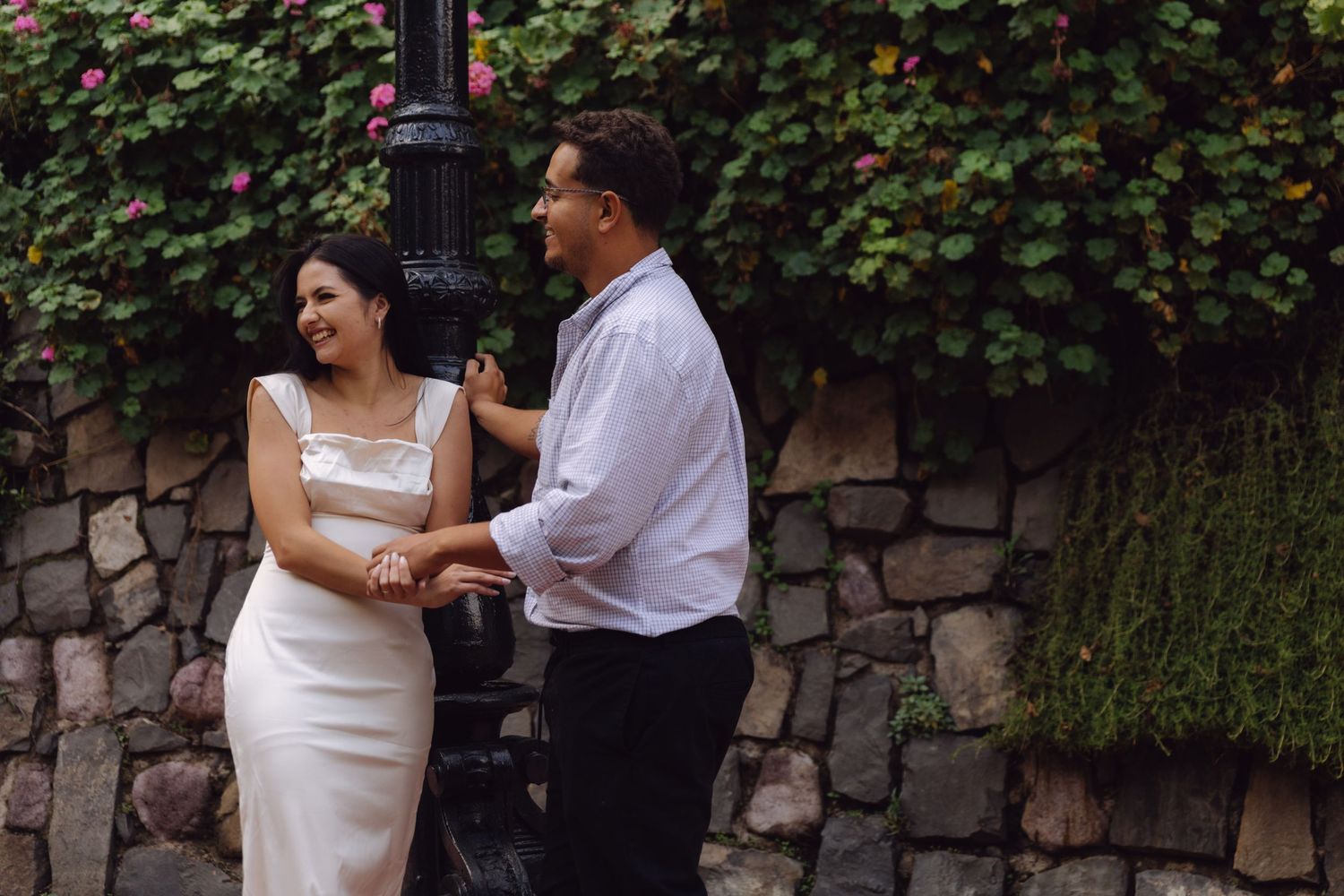 Happy couple walking hand in hand beside ornate lamppost with ivy-covered stone wall.