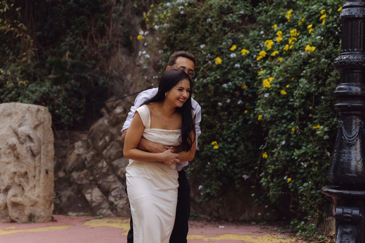 Joyful couple embracing and laughing on pink pathway surrounded by lush garden greenery.