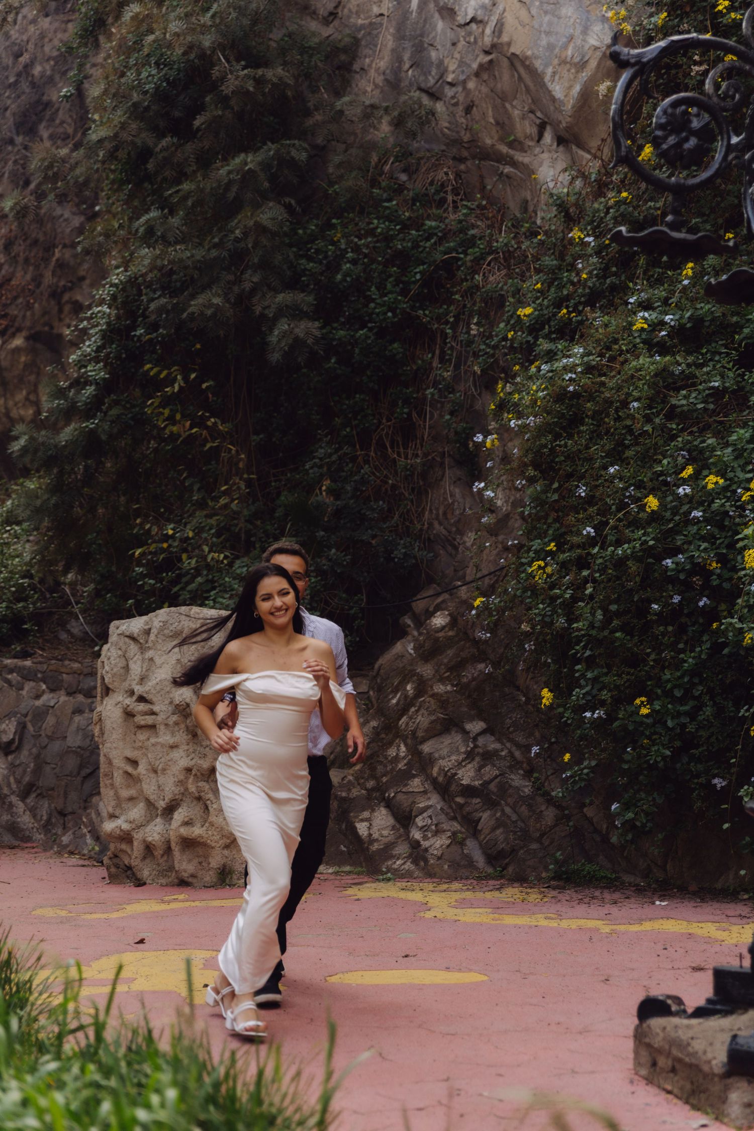 Couple running together on pink pathway with lush green trees and stone wall behind.