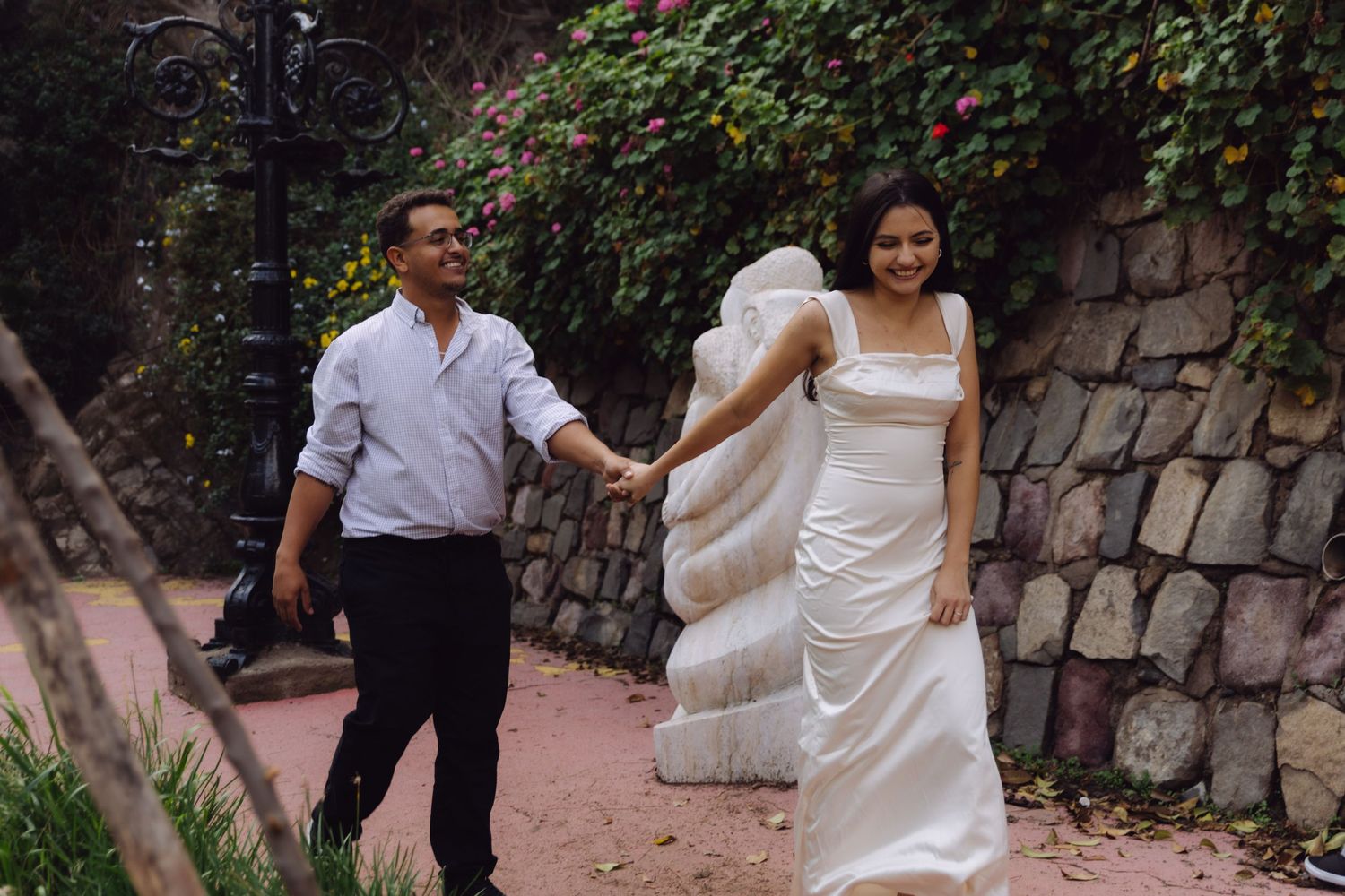 Smiling bride in white dress leading groom along garden path with stone wall backdrop.