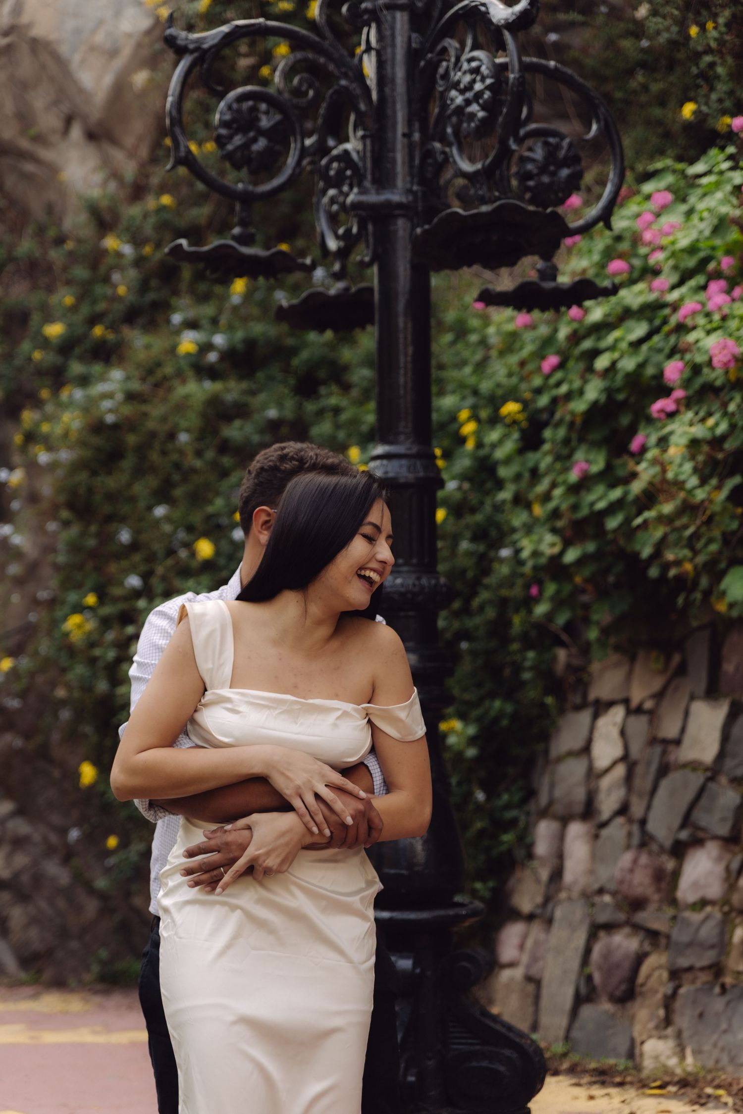 Couple sharing tender embrace by ornate black lamppost with pink roses and stone wall.
