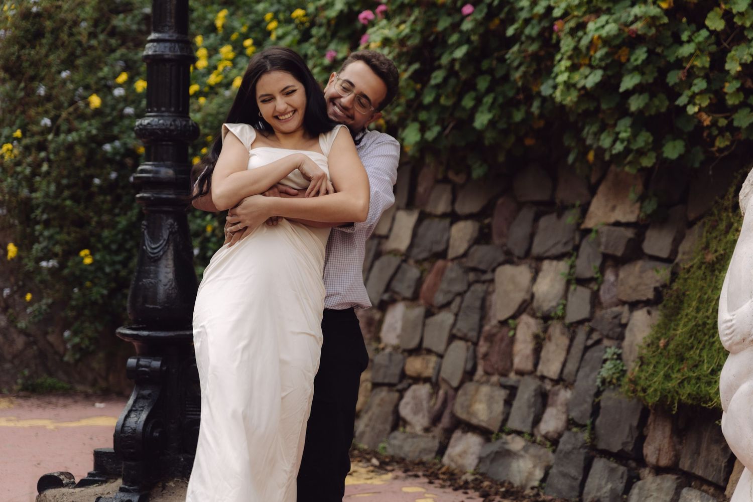 Laughing couple hugging beside black vintage lamppost in lush green garden setting.