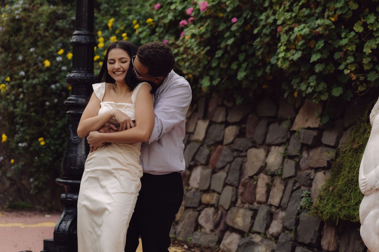 Man kissing womans cheek as she smiles beside lamppost with flowering garden backdrop.