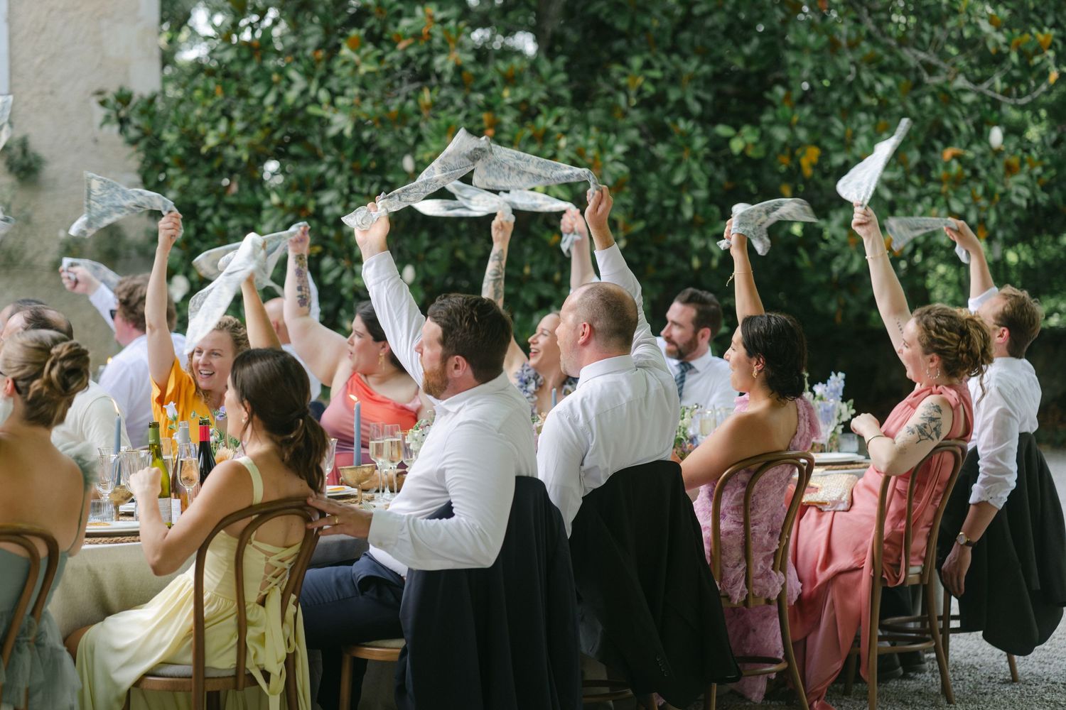 Wedding guests raising paper fans and napkins in celebration at an outdoor reception surrounded by lush greenery.