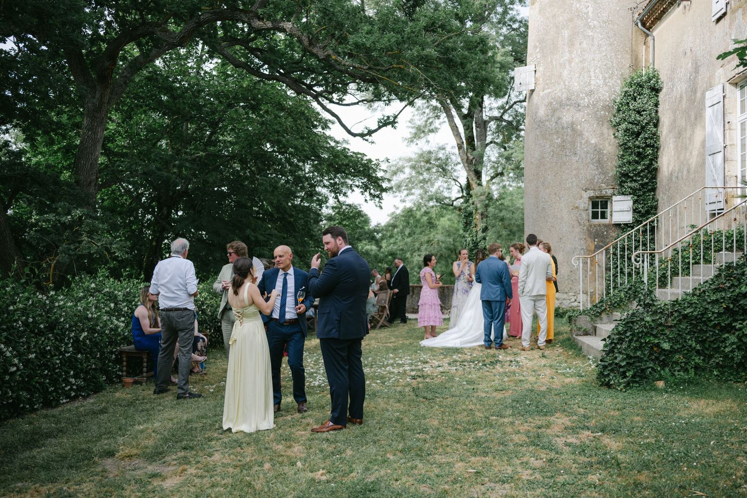 Outdoor wedding reception guests mingling on green lawn beside historic stone building surrounded by lush trees.