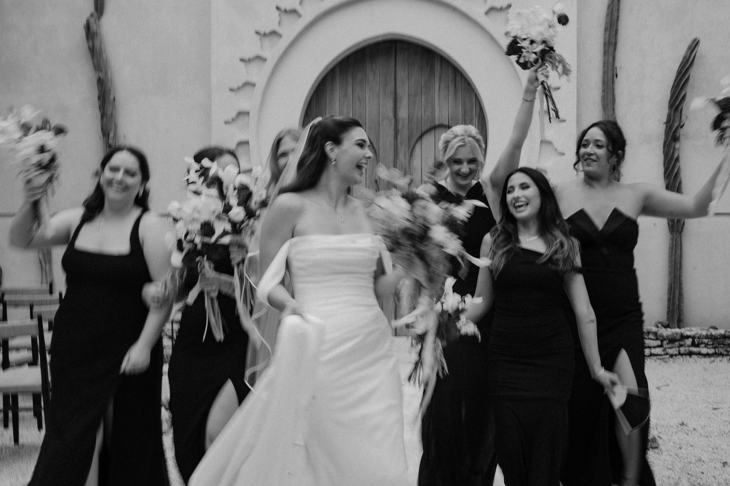 Black and white photo of a bride in a strapless gown walking joyfully with bridesmaids in black dresses holding bouquets.