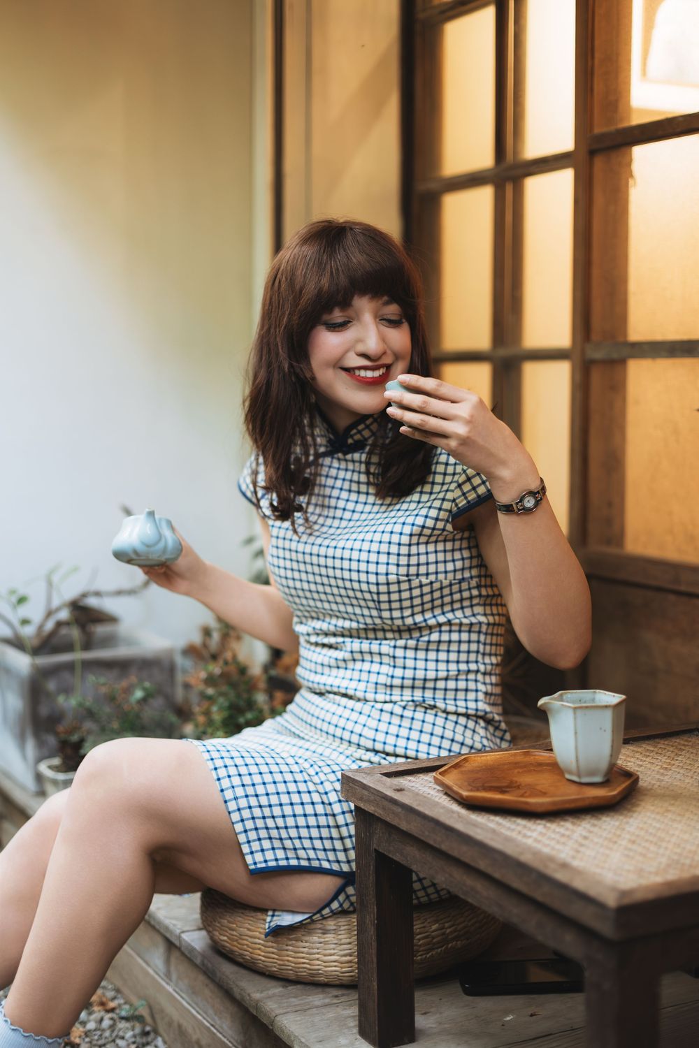 Young woman in checkered dress enjoying tea ceremony outdoors, smiling while holding teacup near wooden table.
