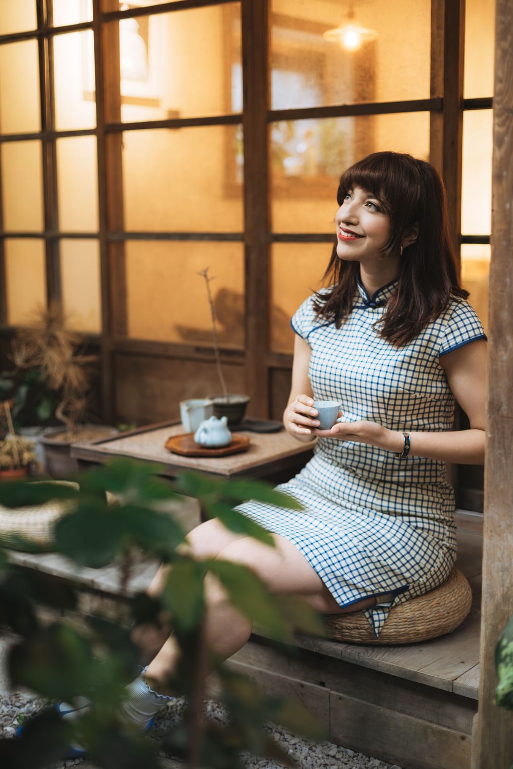 Woman in checkered qipao dress sitting on wooden steps, holding tea cup in a cozy Japanese-style courtyard setting.