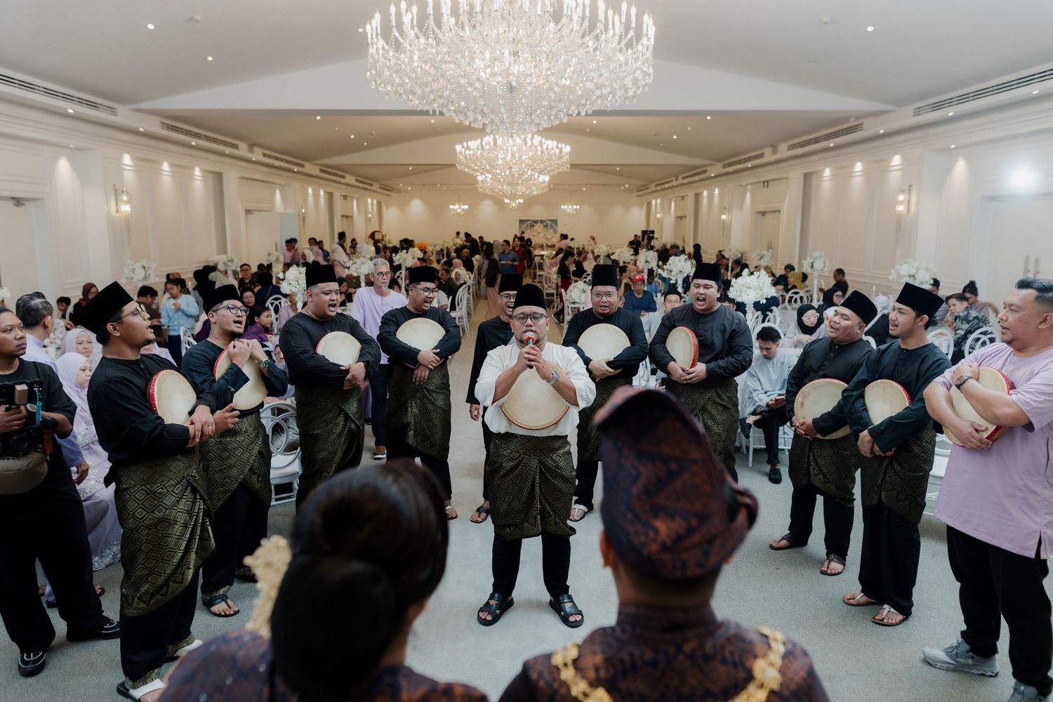 Traditional Malay wedding ceremony with couple seated on stage, performers in green, floral decor, and waterfront backdrop.
