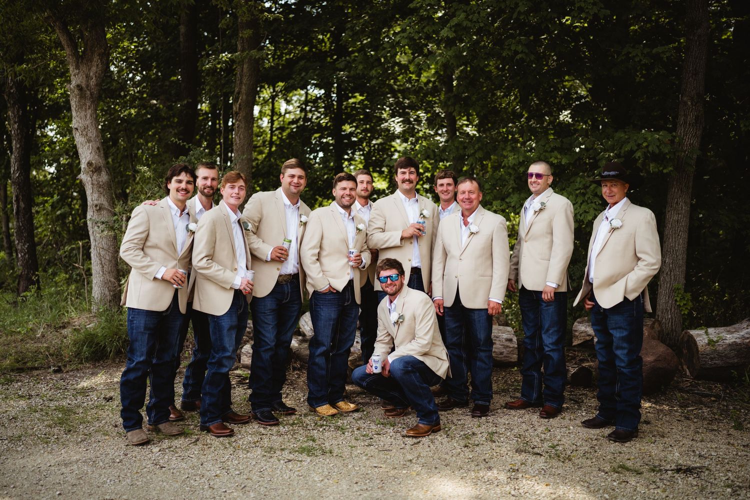 Groomsmen in tan blazers and jeans pose outdoors near trees for a rustic wedding party photo.