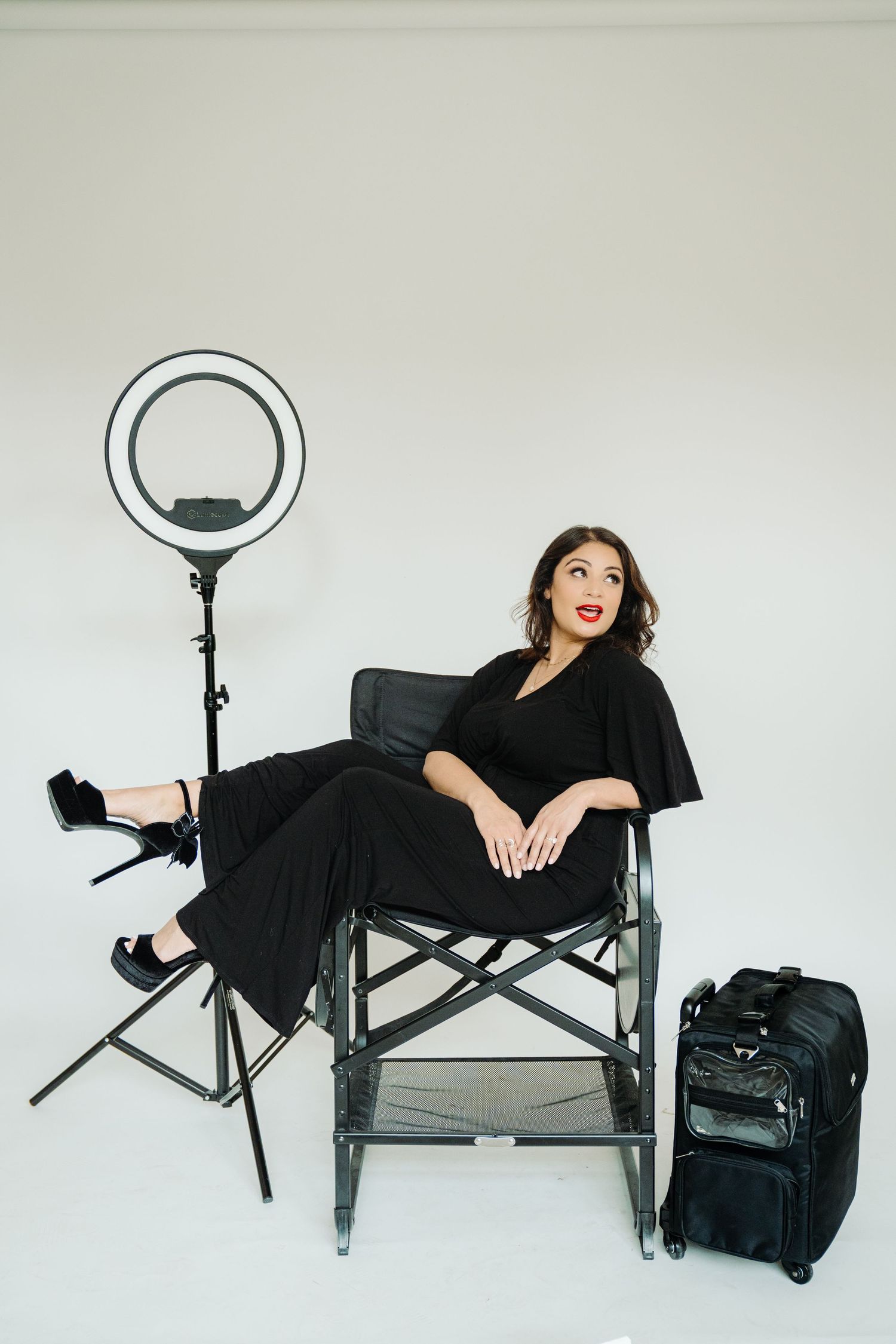 Woman in black outfit sits in director's chair beside ring light and luggage in white studio setting.