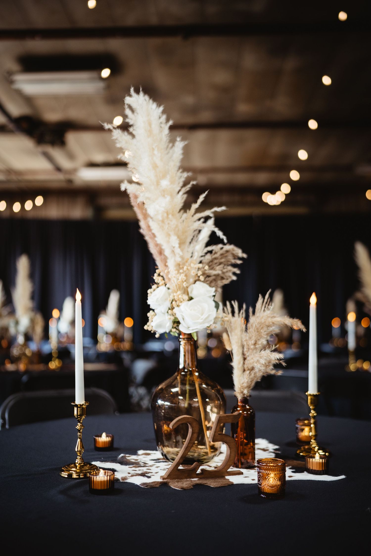 Elegant wedding reception table with pampas grass centerpiece, white roses, gold candlesticks, and glowing candles on black linen.