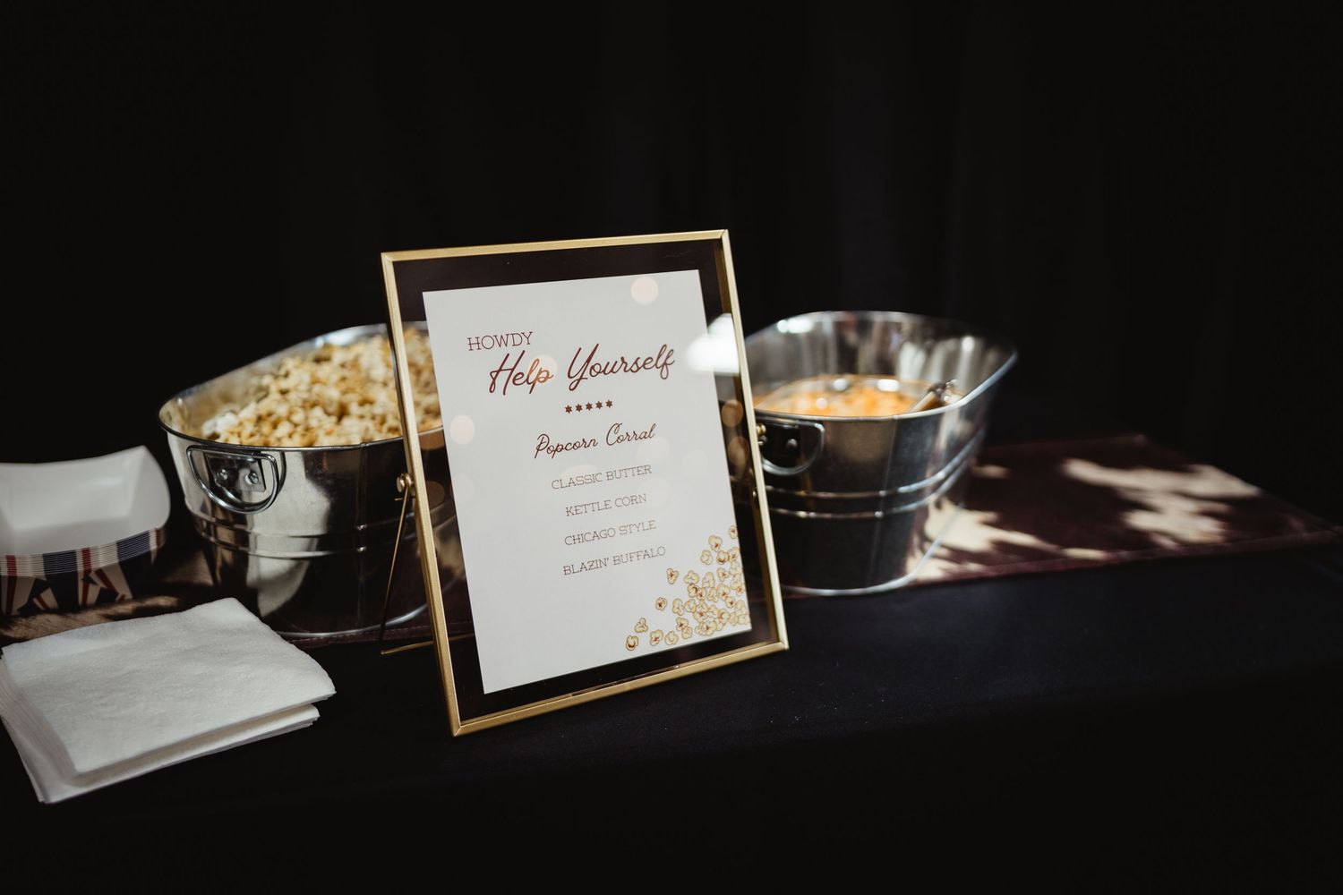 Gold-framed popcorn bar sign on black table with silver buckets at an elegant event.
