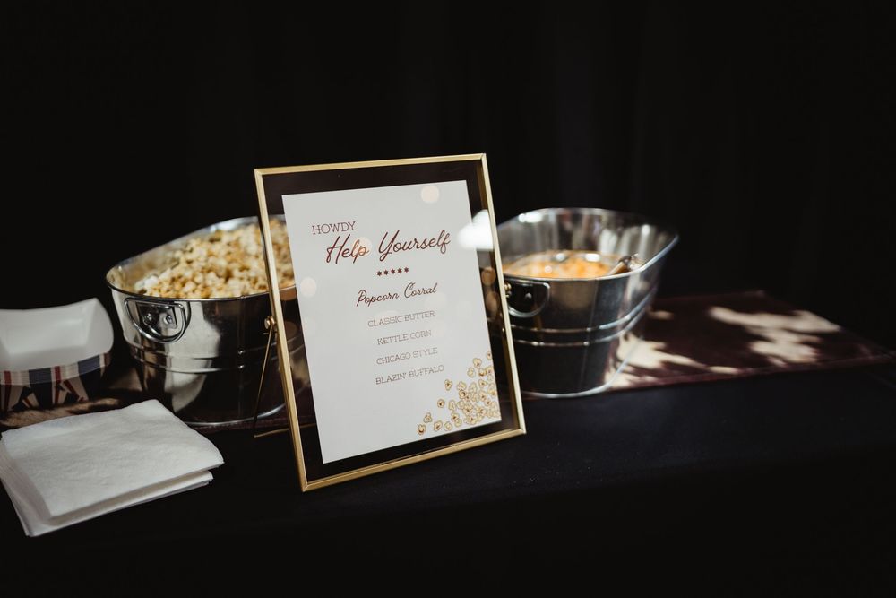 Gold-framed popcorn bar sign on black table with silver buckets at an elegant event.