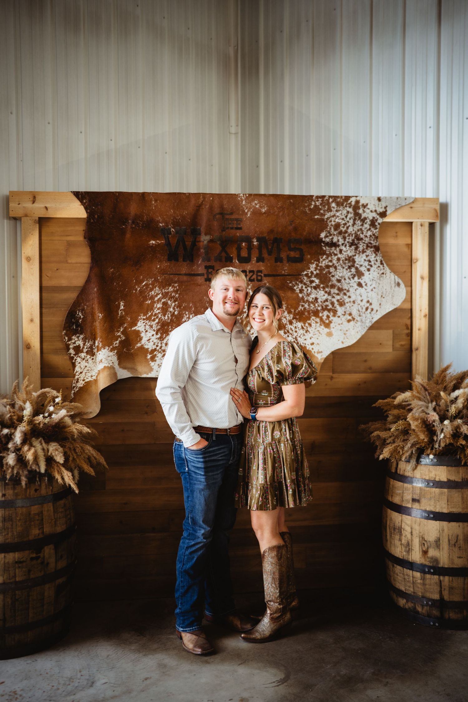 Couple posing in front of rustic wooden backdrop with cowhide sign and barrels in a country barn setting.